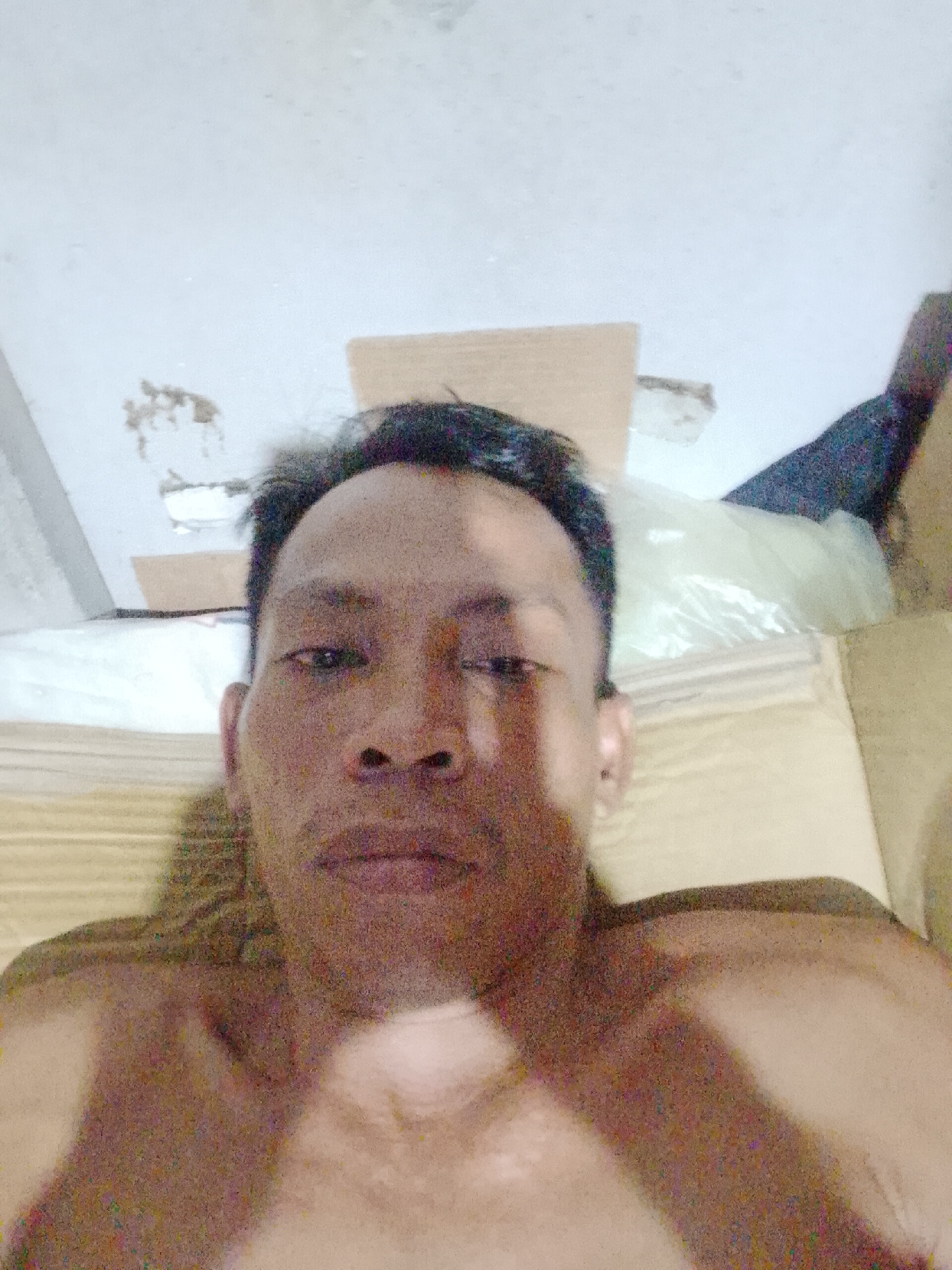 A shirtless man is lying down, appearing to take a selfie. He has dark hair and a weathered complexion. He is situated on a tan-colored surface that appears to be a makeshift bed on the floor. In the background, there are cardboard boxes, a white plastic bag, and what looks like the leg of a dark-colored pair of pants. The location is indicated as Bukit Ketok, Indonesia. The ceiling is white and shows signs of wear.