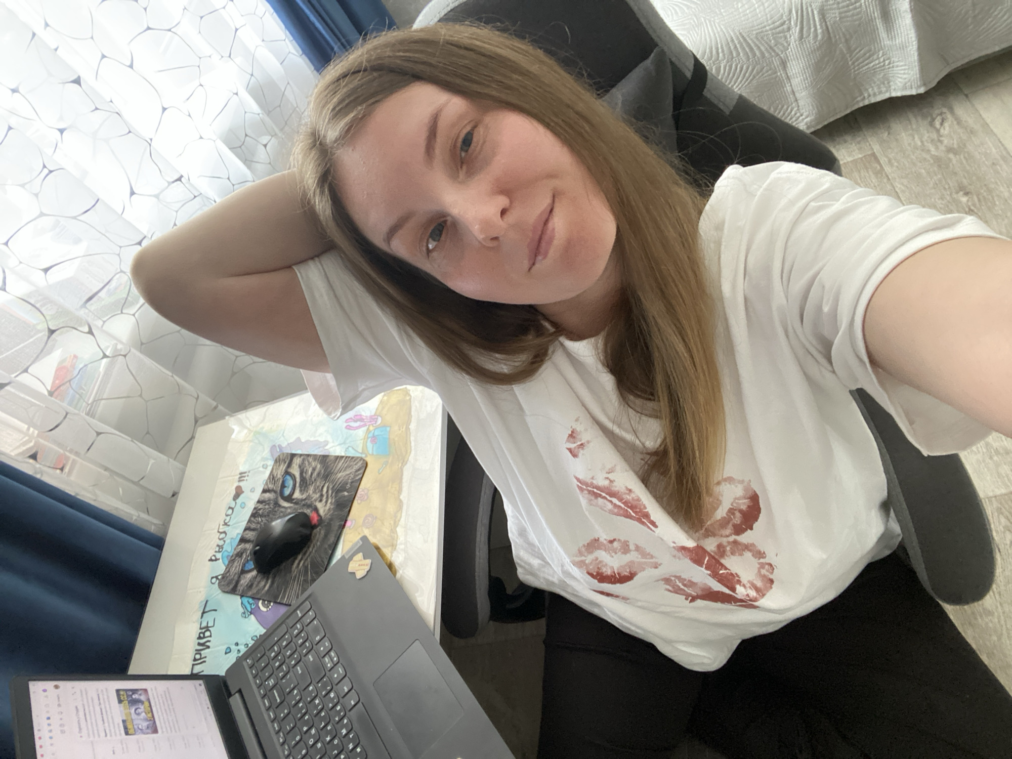 A woman is taking a selfie indoors. She is fair-skinned with light brown hair. She is wearing a white t-shirt with red lipstick kiss marks and black pants. She is sitting in a grey office chair. To her left is a desk with a laptop, a computer mouse on a mousepad depicting a cat, and a drawing or painting. Behind her is a white curtain with a blue pattern and a white bed or sofa. The location appears to be an apartment or home office. There is no indication of the city of Noviy, Russia.