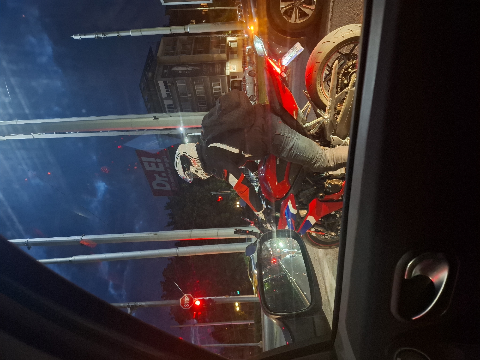 An exterior, eye-level, full shot shows a motorcyclist in a red, white, and black jacket and blue jeans wearing a white helmet, riding a red sportbike. The motorcyclist is facing away from the camera and is viewed from the interior of a car. The motorcyclist is waiting at a red traffic light. The scene takes place at dusk in an urban environment with tall, white poles and a building visible in the background. The sky is dark blue with some clouds. There are other vehicles and streetlights visible.