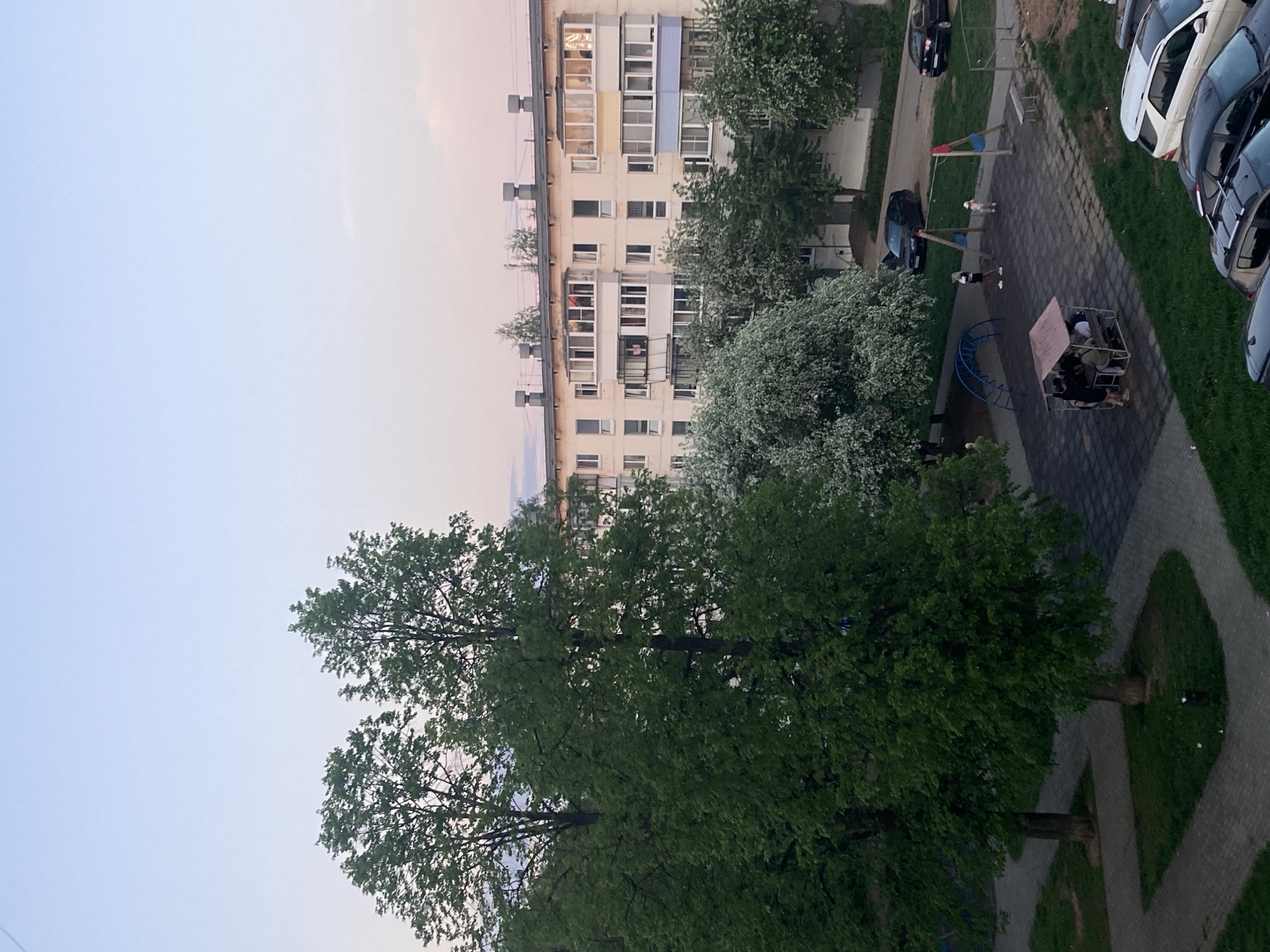 The image shows an aerial view of a residential area in Chaikovsky, Russia. The scene includes a multi-story apartment building with balconies, a playground, a parking lot with several cars, a sidewalk, trees, and patches of grass. People are visible on the playground. The sky is light and colored with hues suggesting early morning or late evening.