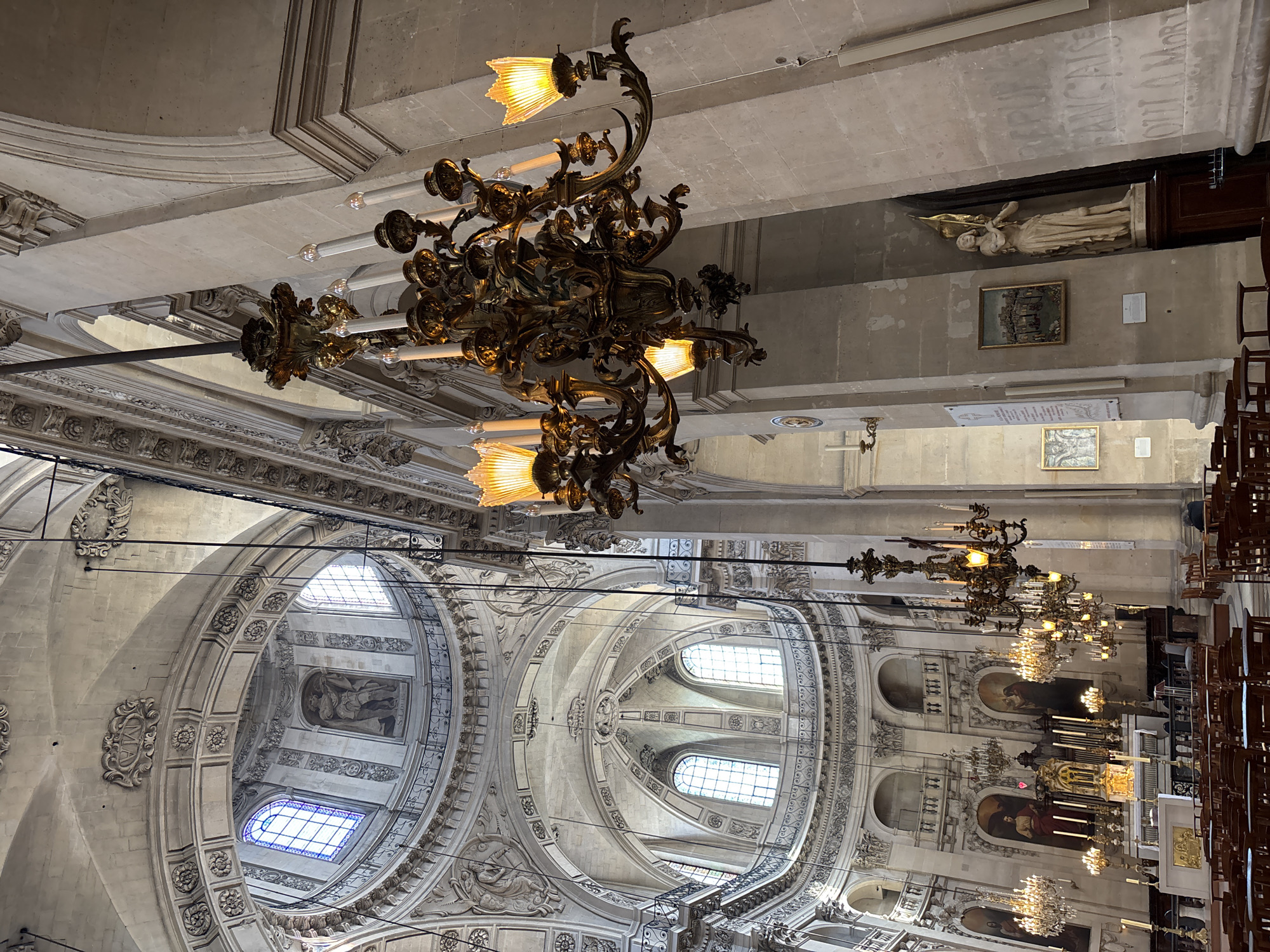 This image depicts the interior of the Église Saint-Louis-des-Invalides in Paris, France. The perspective is from the nave, looking towards the altar and the dome. The scene is dominated by the ornate architecture of the church, with a grand dome visible in the background, adorned with paintings and stained-glass windows. Elaborate chandeliers hang from the high ceilings, illuminated with warm light. Rows of wooden chairs are arranged in the foreground, suggesting a place of worship or a tourist attraction. Paintings and sculptures are visible on the walls and in niches, adding to the rich historical and artistic ambiance of the location. The lighting is a mix of natural light filtering through the windows and the artificial light from the chandeliers, creating a dramatic and awe-inspiring atmosphere.