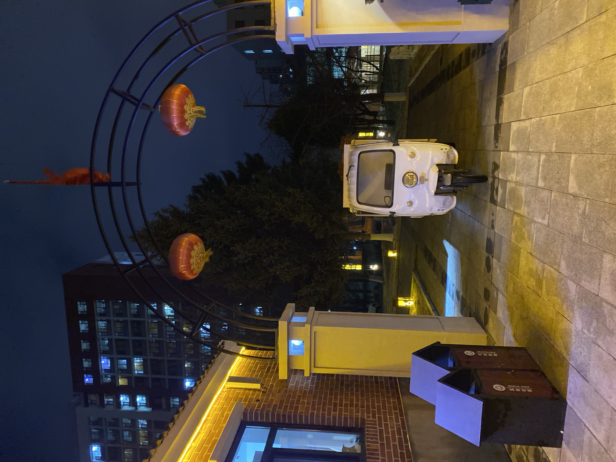 A nighttime view in Zhongtun, China, looking through an archway decorated with red lanterns. A white three-wheeled vehicle is parked beyond the archway. To the right is a paved walkway. In the foreground, there is a building with brick walls and a window. Next to the building, there are two recycling bins labeled in Chinese. In the background is a tall building with many illuminated windows. The sky is dark.