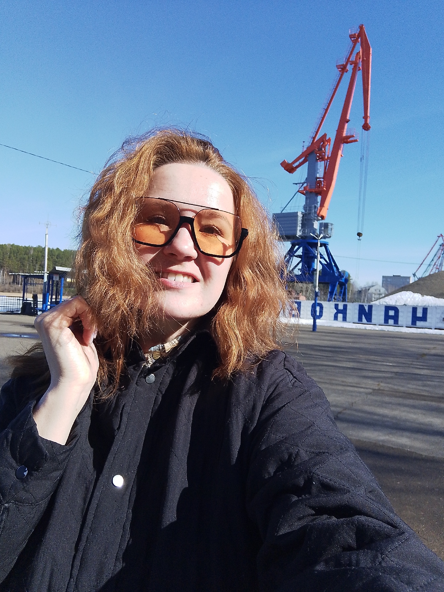 A reddish-haired person, wearing a dark quilted jacket and glasses with orange lenses, is taking a selfie outdoors. The location appears to be an industrial area or port. In the background, there is a large crane painted blue and orange. Behind that is a wall with white lettering that reads "O H N K O H". The sky is blue. The person is smiling, and the overall lighting suggests it is daytime. A tree line is visible in the distance, to the left.