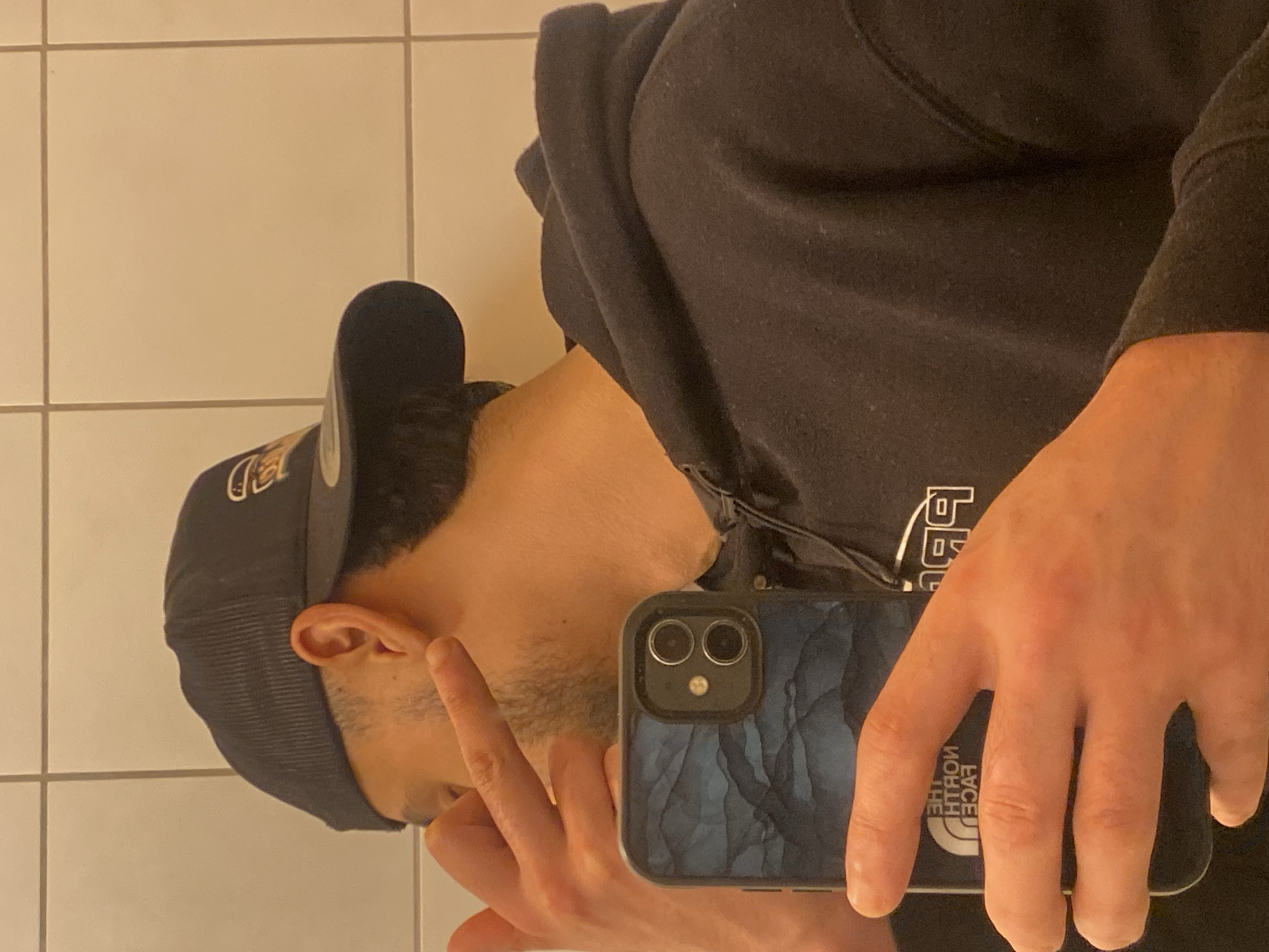 The scene depicts an indoor mirror selfie taken in Schiltigheim, France. A person is visible from the right profile and upper torso, wearing a dark blue corduroy baseball cap with a light-colored logo and a dark, likely black, hooded sweatshirt. Their right hand holds a dark-colored smartphone, featuring two rear camera lenses and a dark blue/gray textured case. Reflected on the phone's screen are reversed text fragments: "THE NORTH FACE" and "99P". The person's left hand is raised with the index finger extended and pointed towards their right ear, performing a gesture. The background consists of a light-colored tiled wall with dark grout lines, indicative of a bathroom or similar utility space. The lighting appears warm and artificial.