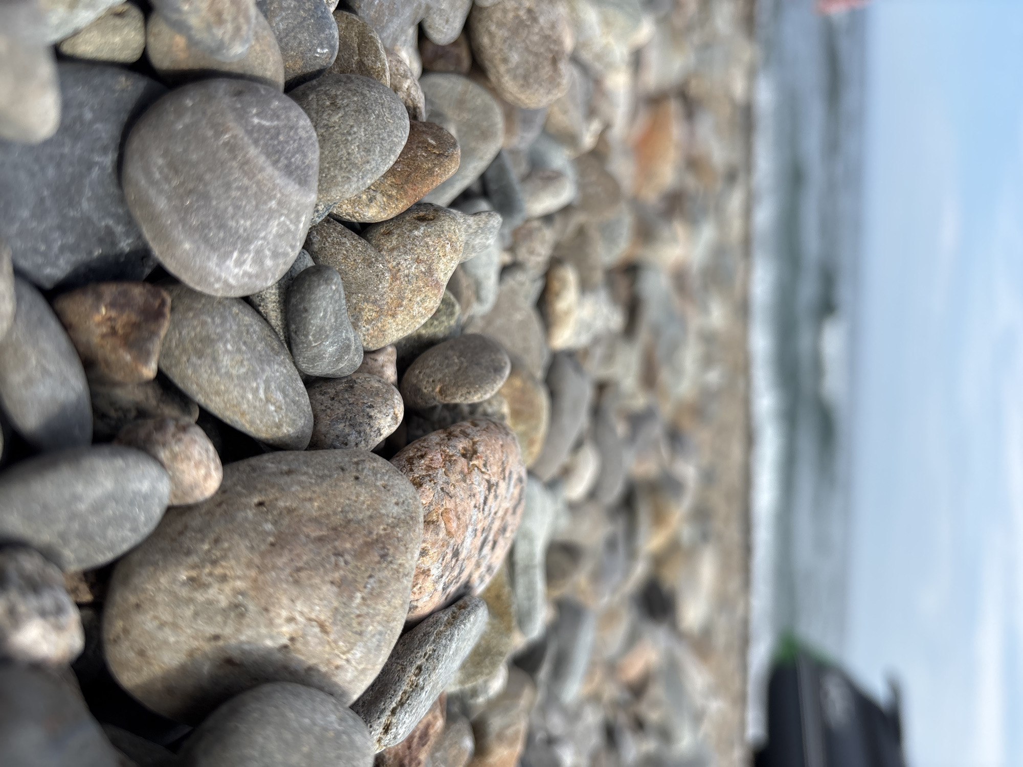 The image is a close-up, low-angle shot of a rocky beach. The foreground is dominated by numerous pebbles and larger stones of various shapes, sizes, and colors, predominantly grays, browns, and some with reddish hues. The stones are smooth and rounded, suggesting they have been shaped by water.
In the background, the ocean is visible with gentle waves breaking on the shore. The water is a muted blue-green. Above the water, the sky is a pale blue with some wispy white clouds.
On the far right of the background, a dark object with a green accent, possibly a jet ski or personal watercraft, is partially visible near the water's edge. A small portion of what appears to be a person's arm, wearing a pink garment, is visible on the far left side of the background, also near the water.
The overall impression is a tranquil scene at a beach. The location is specified as Rechka Mishikha, Russia.