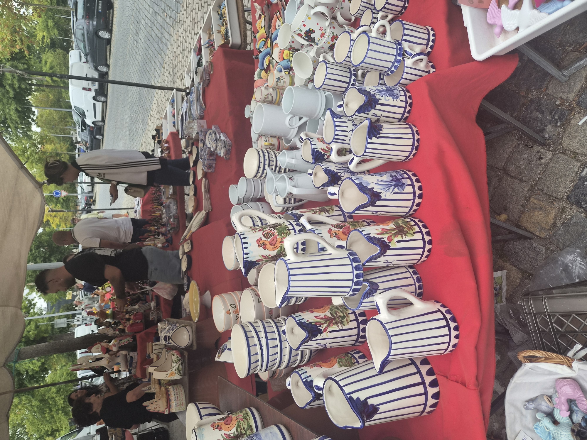 This is a street market scene in Barcelos, Portugal, featuring a stall laden with ceramic tableware, primarily jugs and bowls. The items are predominantly white with blue stripes and rooster motifs, characteristic of Barcelos pottery. Several people are visible browsing or attending the stall. In the background, parked cars and a cobblestone street indicate an urban setting. The market is set up under a tent, offering shade. The overall atmosphere suggests a lively daytime event.