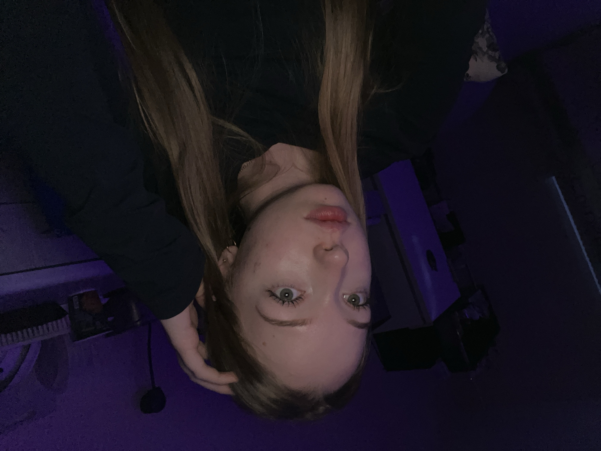 A young woman with long brown hair is positioned upside down in the frame. She is wearing a black top and is touching her hair with her left hand. Her face is visible, and she has light skin, green eyes, and reddish lips. The background is dark with hints of purple lighting. There are indistinct shapes and objects in the background, including what appears to be furniture or equipment. The location in Chaikovsky, Russia is not discernible from the image.