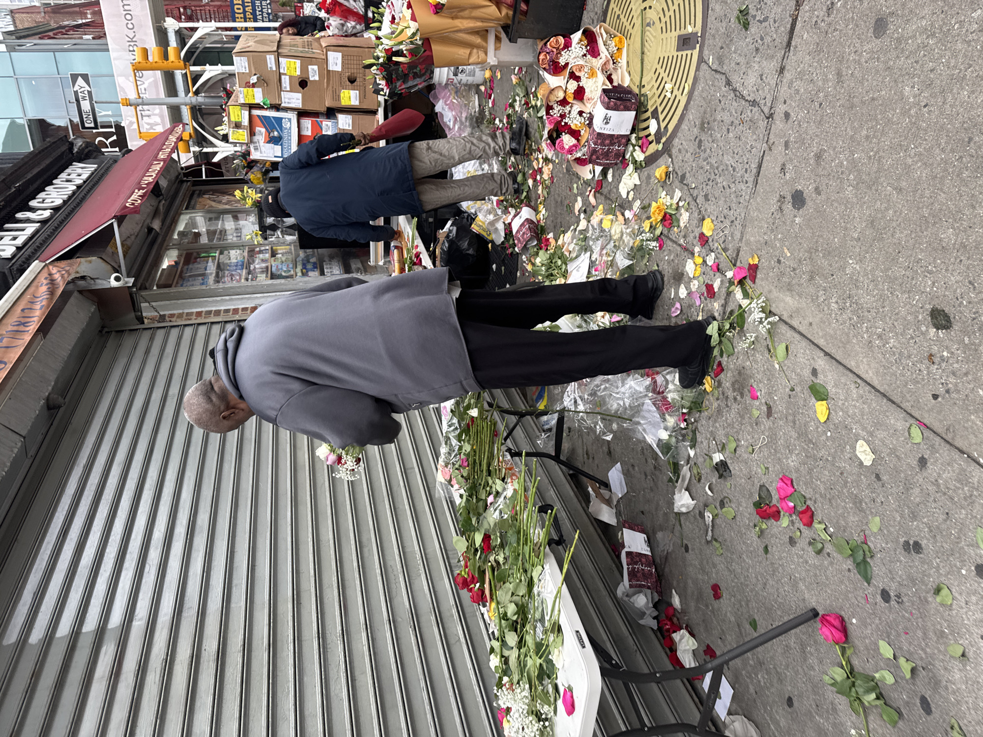 The image shows the exterior of a store in New York City, United States of America. The store has a red awning with "BELL & GROCERY" printed on it, and a sign above that reads "COPE ЧАМОИИ НІТЫЛЛА THEVEBK.com". A metal security shutter is pulled down over the store's front.
Two people are visible in the image. One is wearing a grey hooded sweatshirt and black pants, and the other is wearing a dark blue jacket and tan pants. They are standing near a makeshift memorial outside the store.
The memorial consists of flowers, including roses, scattered on the sidewalk. There are also some boxes and bags present, along with a black trash bag. A yellow traffic light pole can be seen in the background, as well as a manhole cover on the sidewalk.
