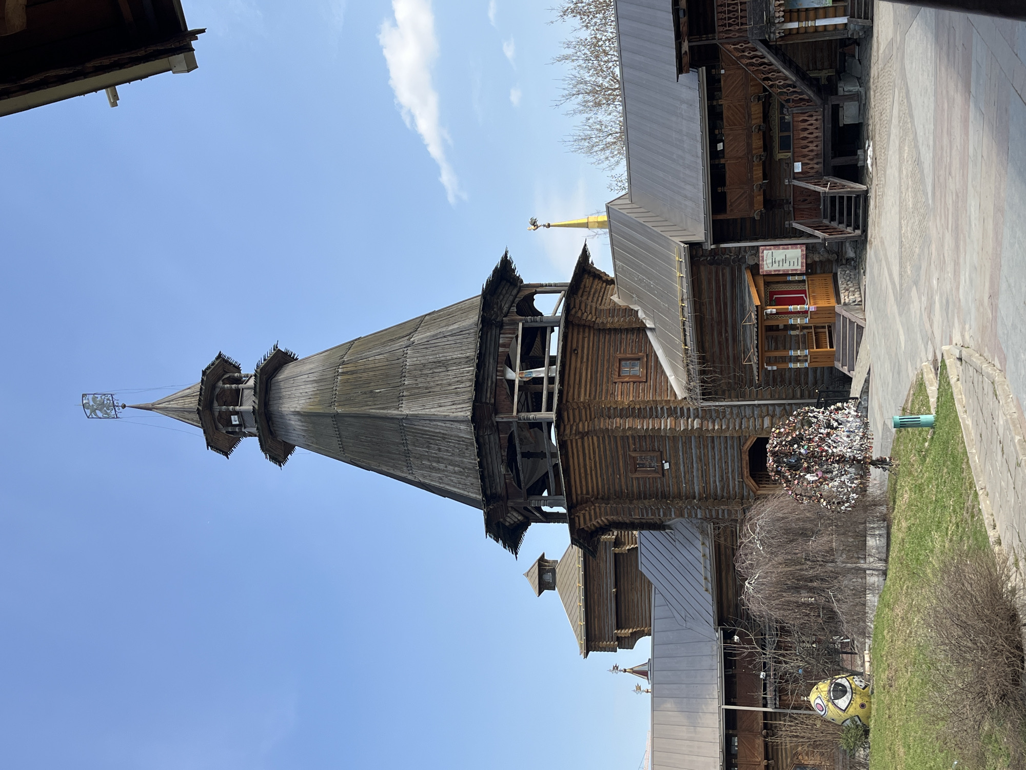 The image shows a tall, wooden structure with a conical roof, likely a church or tower, in Moscow, Russia. The structure is multi-tiered, with visible wooden details and ornamentation. Adjacent to the structure is another building with a gray roof, possibly connected or a part of the same complex. The sky is clear and blue with a few clouds. There is landscaping including patches of grass and a decorative tree covered in locks. The ground is paved. A large rock painted as a minion can be seen next to the landscaping.