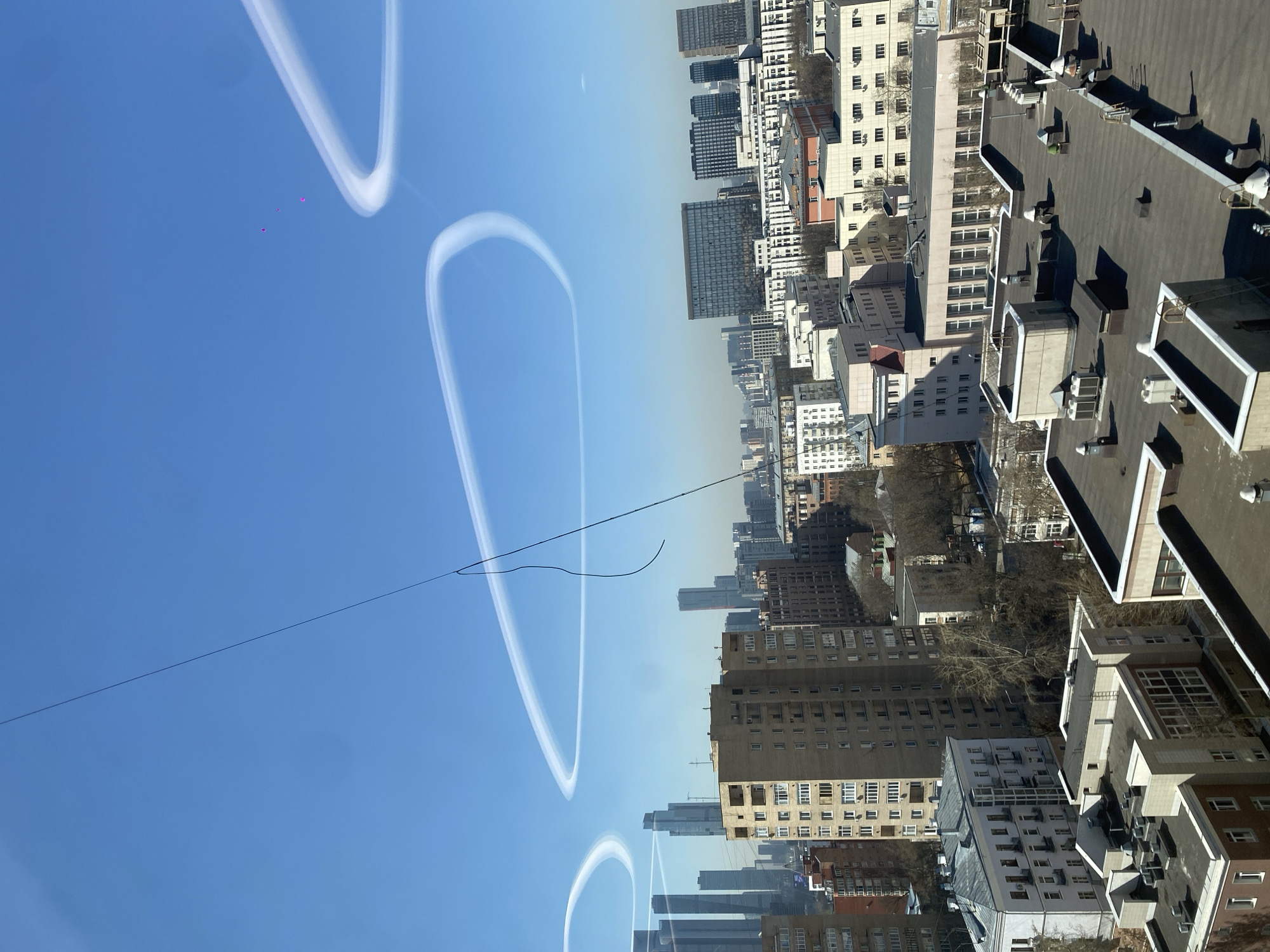 The image presents a cityscape, likely Moscow, Russia, under a clear blue sky with contrails forming distinct circular patterns. A thin, dark wire or cable stretches across the sky. The buildings are a mix of mid-rise and high-rise structures, varying in color from light to dark, with visible architectural details. Rooftops are diverse, featuring both flat and slightly pitched surfaces, some with visible ventilation or HVAC systems. Patches of trees, bare of leaves, are visible interspersed among the buildings. The image is slightly rotated, giving a tilted perspective of the horizon. There are some tiny pink objects in the sky.