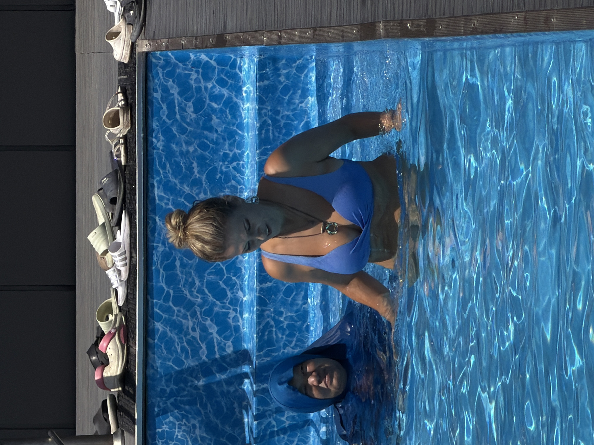Two individuals are in a swimming pool. The individual on the left is wearing a blue full-body covering, with their face partially visible. The individual on the right is wearing a blue bikini and a necklace. They are both submerged in blue water, with stairs visible in the background. Above the pool, a collection of footwear, including sandals and flip-flops, is arranged on a dark surface. The environment appears to be outdoors, possibly a resort or a private residence.