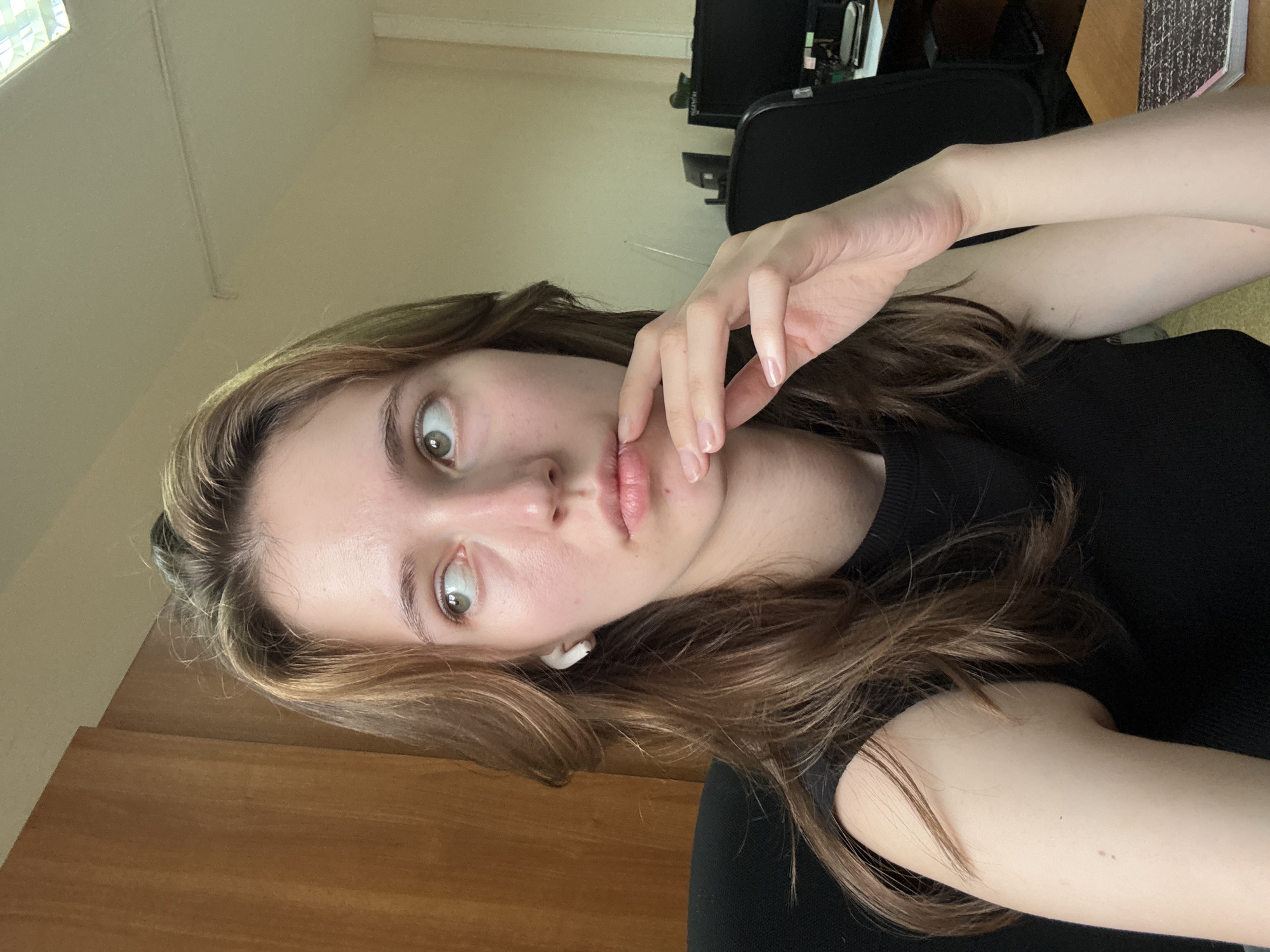 The media file shows a close-up, head-and-shoulders shot of a young woman with light brown hair. She is wearing a black top and white wireless earbuds. Her left hand is resting on her chin, and her eyes are wide open, looking directly at the camera. Her lips are slightly parted. The lighting suggests it might be daytime, with natural light coming from a window, possibly a home or office setting. In the background, there are hints of office furniture, including a black chair and a computer monitor, suggesting a work or study environment. The image appears to be a selfie, taken from a slightly angled perspective. The woman's expression is one of surprise or curiosity. There is no visible text in the image. The location context is Perm, Russia.