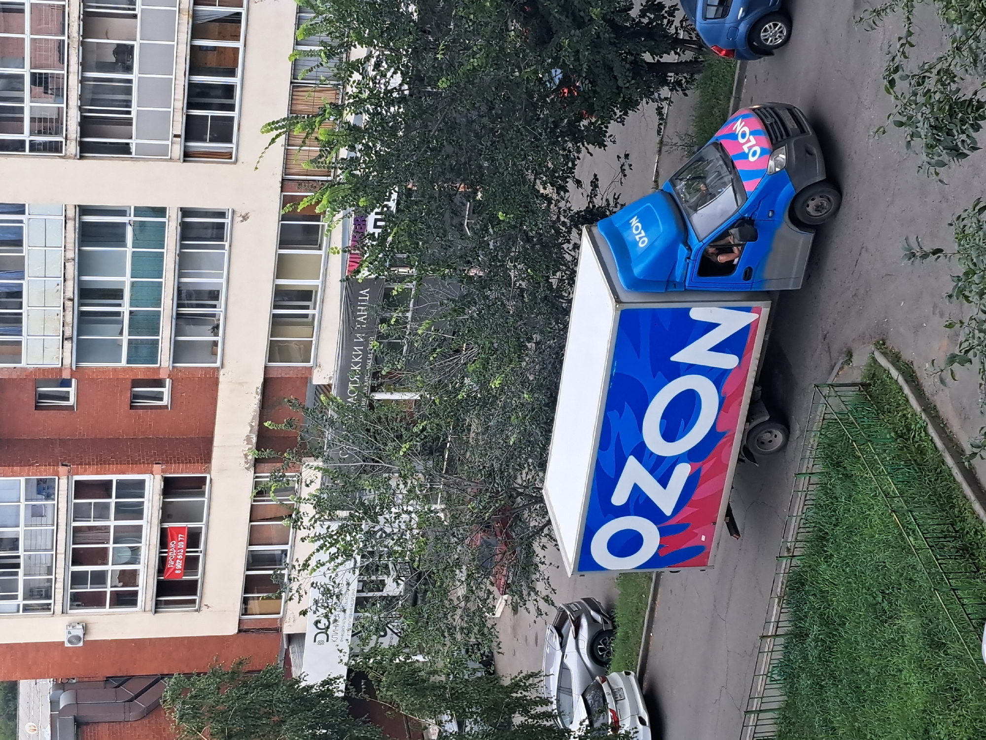 An Ozon delivery truck with its branding is driving on a paved road in a city. The truck is blue with the Ozon logo prominently displayed in white, pink, and blue. The side of the truck has large white letters that spell "OZON" with a wave-like pink and blue design. The truck has a blue cab with a driver visible inside. The road surface is asphalt with visible cracks. To the left of the truck, there are several parked cars, including a silver SUV. In the foreground, there is a green, fenced grassy area. Behind the road and cars, there is a multi-story brick and cream-colored residential building with many windows, some of which have balconies or enclosed glass areas. Some windows have red banners hanging from them. Trees with green foliage are also visible in the scene, obscuring parts of the building and the background. The image appears to have been taken from an elevated perspective, looking down on the street.