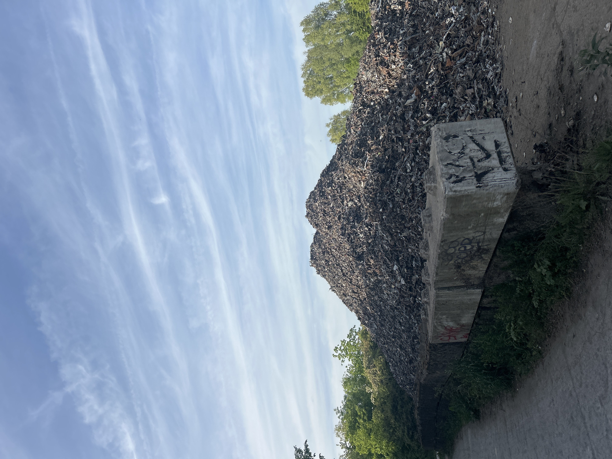 The image shows a large heap of scrap metal or waste material adjacent to a concrete barrier. The barrier appears to be marked with graffiti. The sky is visible with streaks of clouds. Vegetation, possibly trees, is visible behind and to the side of the scrap pile. The scene is located in Marquette-lez-Lille, France. No people are visible. A grey ground or pavement is in the foreground of the photo.