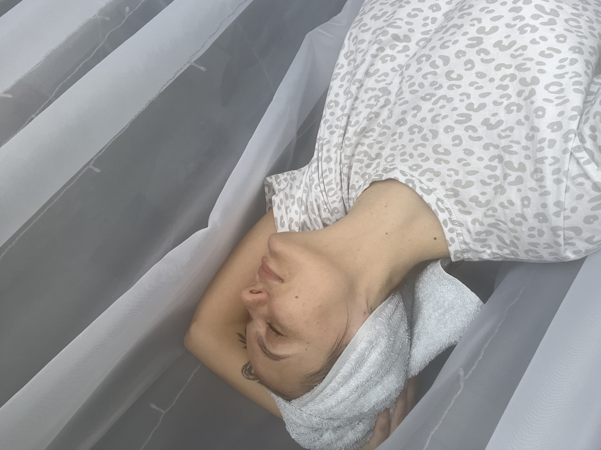 A person is lying down with a towel wrapped around their head. They are wearing a light-colored shirt with a leopard print pattern. The person appears to be resting or undergoing a spa treatment. The background consists of draped, sheer white fabric, possibly a curtain or canopy. The location is described as the city of Chaikovsky, Russia.