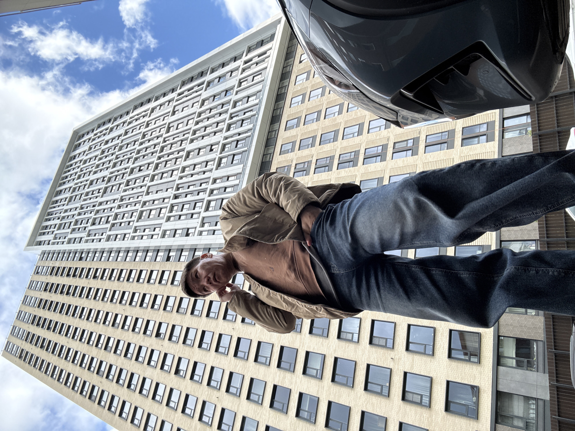 A low-angle shot depicts a man in a brown shirt, tan jacket, and dark jeans, appearing to look down at the camera. Above him are two tall buildings. One is light tan with many dark, rectangular windows. The other is white with a similar pattern of dark, rectangular windows. A portion of a dark-colored car is in the top right corner. The sky is visible in the top left with patches of blue and white clouds. There's no verifiable information to indicate the location is Khimki, Russia.
