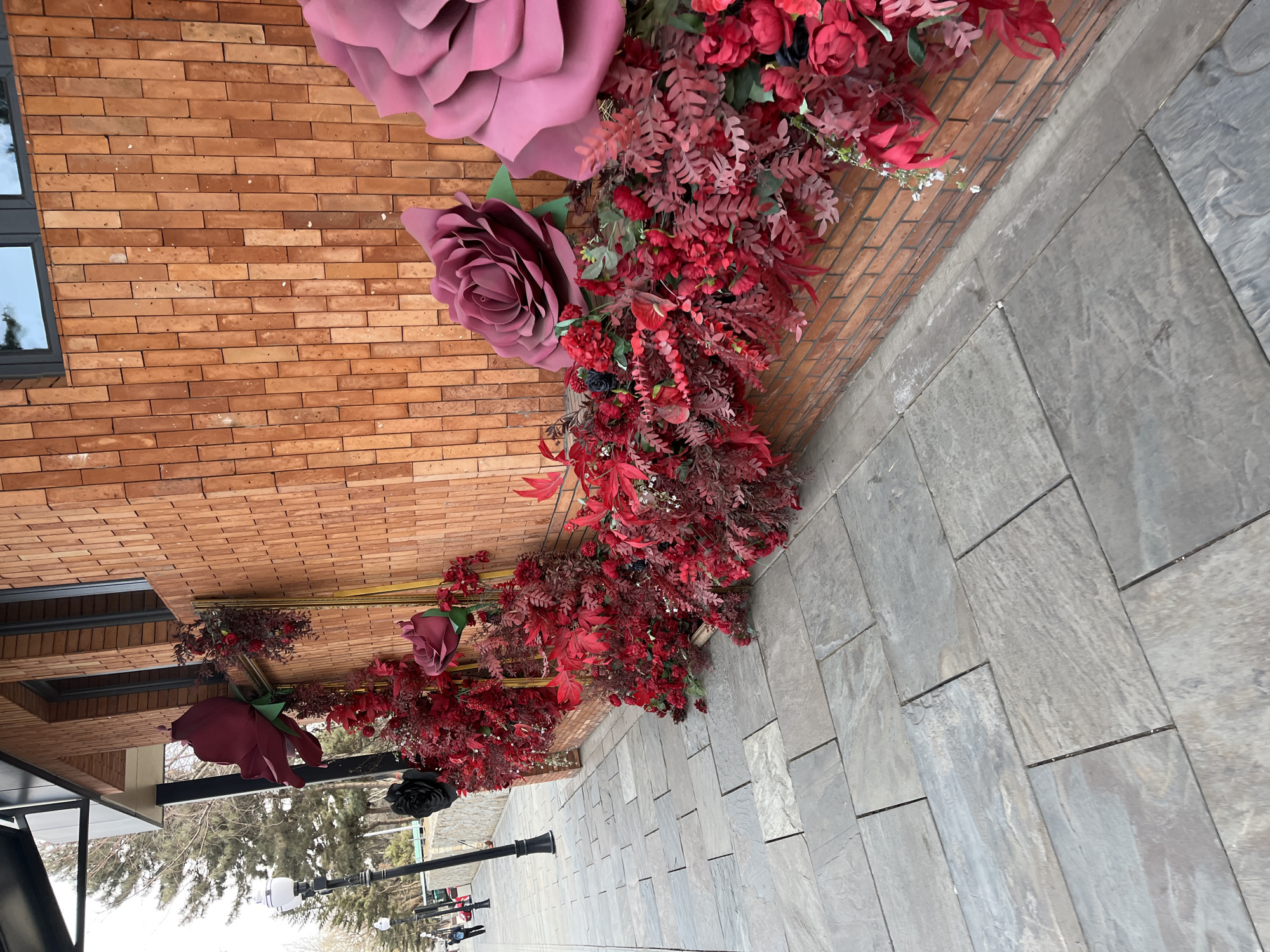 The image shows an outdoor setting. A brick wall is adorned with large artificial flowers, primarily roses in shades of red and pink. The flowers and accompanying greenery are arranged in a garland-like fashion along the wall. In the foreground, a paved walkway stretches into the distance. Street lamps are visible along the walkway, and beyond them, trees and buildings can be seen. Based on visual cues, the location is consistent with a modern, possibly commercial area in an urban environment. Further forensic details would require additional context or higher resolution for identifying specific features. Given the urban setting with a mix of greenery and modern architecture, this could be a park or pedestrian area in Changchun City, China. However, without additional specific landmarks or details, this remains a general assessment.