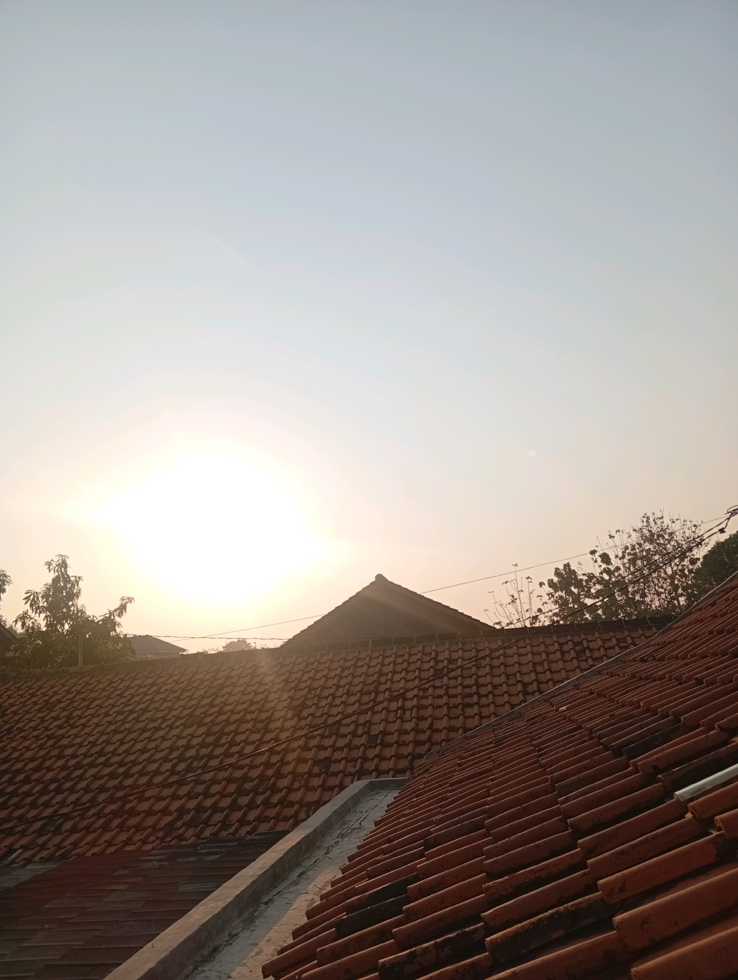 The image captures a view of rooftops in Kebandingan, Indonesia, during the late afternoon or early morning, evidenced by the low-angled sunlight. The sky is a soft gradient of pale blue and white, with the sun appearing as a bright, almost blown-out orb in the upper left quadrant. Terracotta tiled roofs dominate the foreground and middle ground, arranged in overlapping patterns. Some tiles appear aged and show signs of wear. The roofs are on residential buildings, indicated by their pitched design. Trees with dense foliage are visible behind the roofs on the left, and sparser, leafless branches are seen on the right, silhouetted against the sky and suggesting a drier season. Power lines are strung between poles in the distance. The overall scene conveys a quiet, domestic atmosphere.