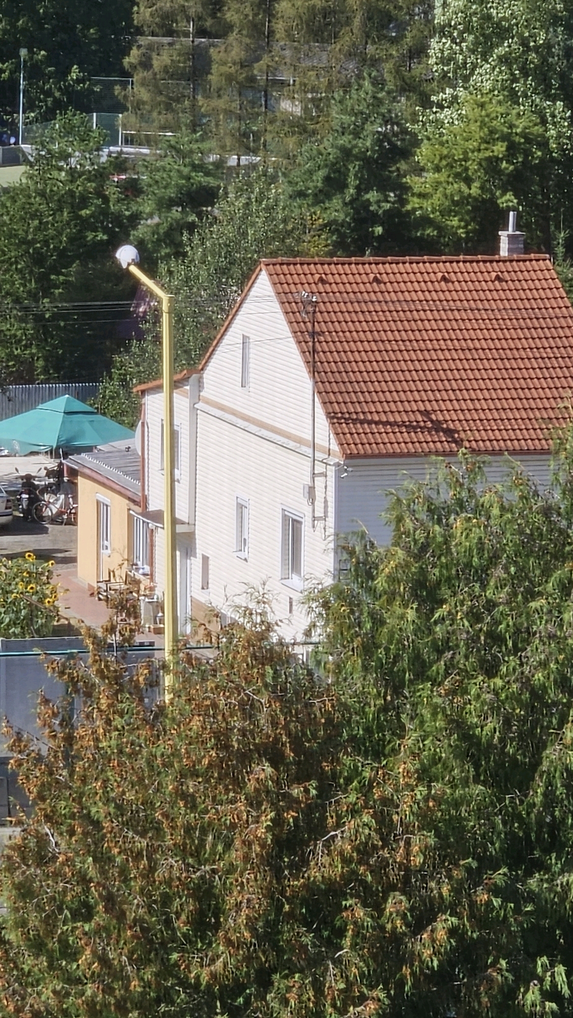 The image depicts a residential area with houses and trees. In the foreground, a large tree with green and brown foliage obscures part of the scene. Behind it, a cream-colored house with a red-tiled roof is visible. To the left of this house, a smaller yellow house with a gray roof is attached. A bright yellow lamppost stands prominently in front of the cream-colored house. In the background, a green tent-like structure is partially visible, along with what appears to be a motorcycle and a bicycle. Further in the background, there are more trees and some fencing, suggesting a park or sports facility. The overall impression is a suburban or semi-rural setting, likely in Prešov, Slovakia, given the context of the request. There are no people clearly visible in the image, and no specific actions or interactions are depicted. The scene appears to be captured during daylight, under sunny conditions.