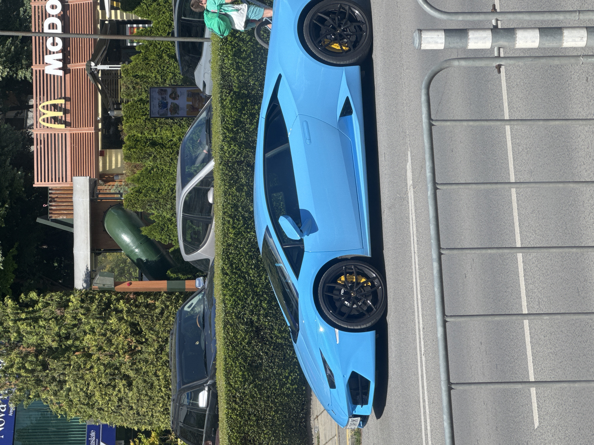 The image shows a blue Lamborghini parked on a street in Varna, Bulgaria. Next to the car is a McDonald's restaurant. A black SUV and a silver car are also parked nearby. A person is visible on a bicycle, near the McDonald's. The street has a metal barrier. There is greenery along the side of the street.