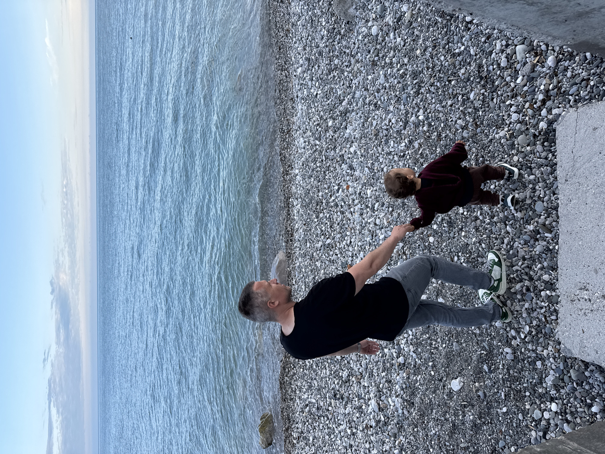A father and child are on a pebble beach in Abaata, Abkhazia, next to the Black Sea. The child, dressed in a maroon long-sleeve shirt and brown pants with dark shoes, is being held by the hand by the father, who is wearing a black shirt, gray pants, and green-accented tennis shoes. The beach is composed of light gray pebbles and is bordered on one side by the sea, and on another by a gray concrete structure. The sky is light blue, and the sea is a matching light blue with gentle ripples.
