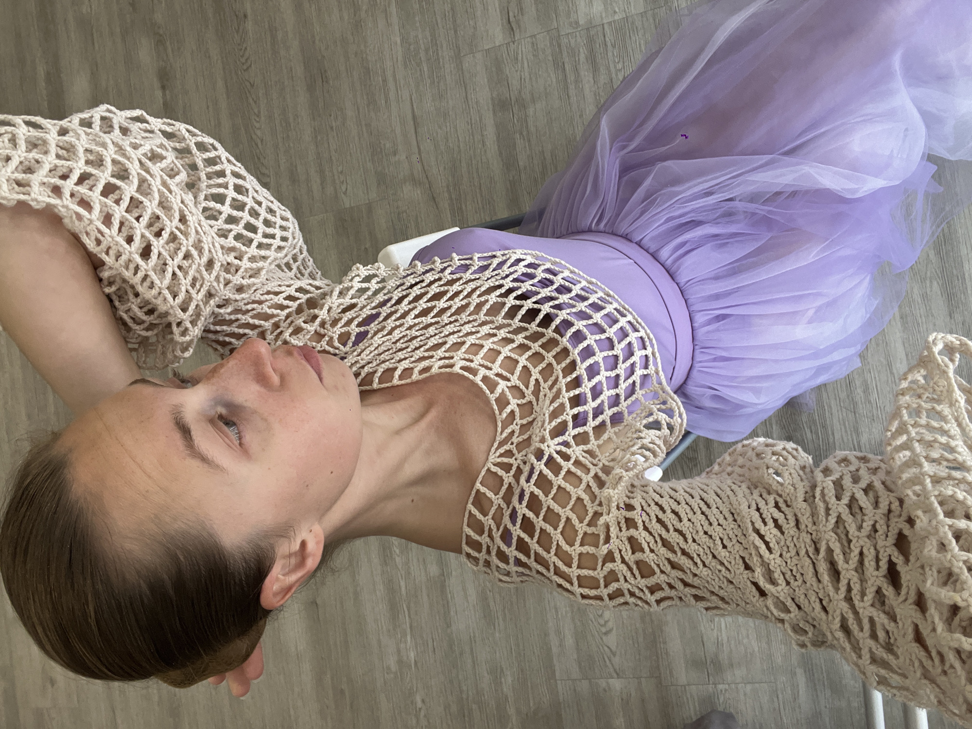 An overhead photograph depicts a seated female individual indoors, located in Budva, Montenegro. Her light brown hair is pulled back into a bun, and her fair skin displays visible freckles on the face and neck. She is dressed in a light purple strapless dress featuring a voluminous tulle skirt, overlaid with a cream-colored, open-weave crocheted top that includes wide sleeves. Her right arm is raised, with the hand positioned near the back of her head. Her gaze is directed towards the right side of the frame. The immediate environment includes a light greyish-brown wooden floor with horizontal planking, and a segment of a white chair back is visible behind her.