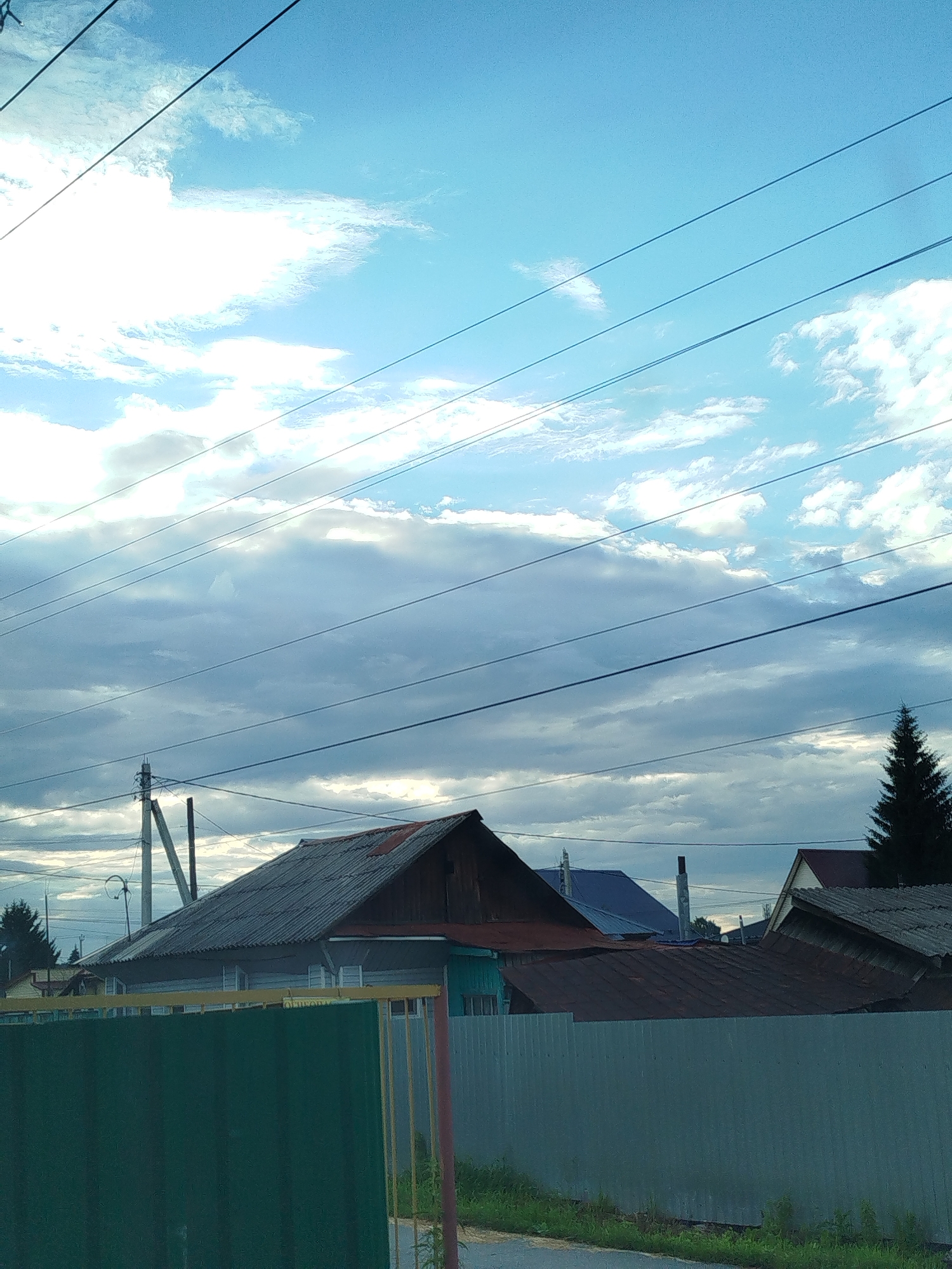 The media file shows a rural landscape with houses, power lines, and a cloudy sky. The scene is set in Zavodoukovsk, Russia. The time of day appears to be late afternoon or early evening, with the sun casting some light through the clouds. The weather is overcast with scattered patches of blue sky.
In the foreground, a green metal fence runs horizontally across the image, partially obscuring the view of the houses behind it. The houses are low-rise buildings with pitched roofs made of corrugated material. Some of the houses appear to be made of wood, while others have siding. A tall power pole with visible wires is situated to the left of the frame, indicating the presence of infrastructure. In the background, a dark evergreen tree stands out against the lighter sky.
There are no people visible in the image. The overall mood of the scene is quiet and somewhat somber, perhaps due to the overcast sky. No text is visible in the media file. The image captures a typical scene in a rural Russian settlement.