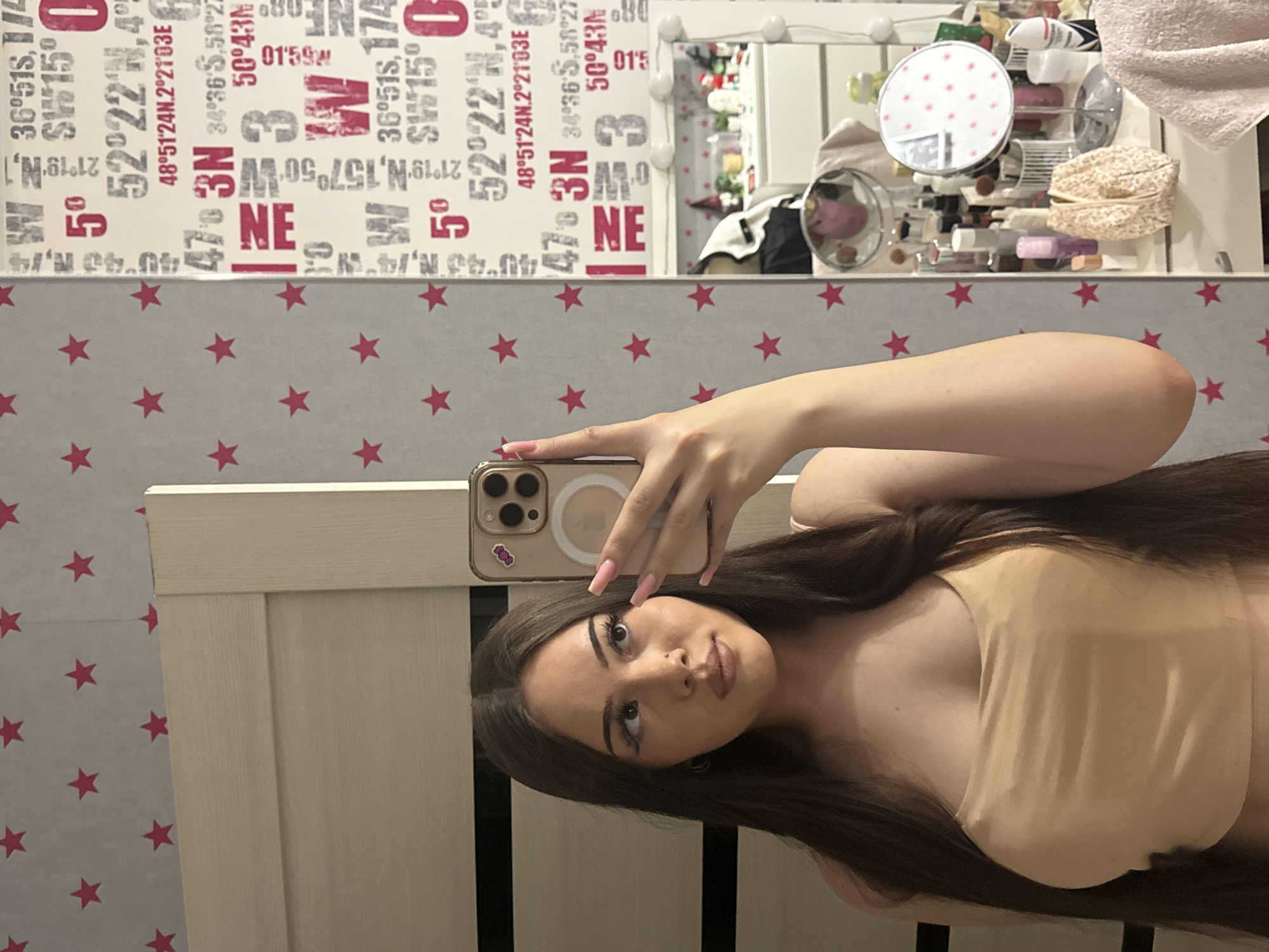 A young woman is captured in a mirror selfie, taken indoors, likely in Naberezhnye Chelny, Russia. She has long, dark brown hair flowing over her shoulders and is wearing a light-colored, strapless top, possibly beige or peach. Her face is turned slightly towards the camera with a neutral to subtle expression, featuring noticeable makeup including eyeliner, lip color, and a small piercing above her upper lip. She holds an iPhone, equipped with a light-colored case and a circular MagSafe accessory, her long, light pink nails clearly visible.
The background wall behind her is divided into two distinct sections. The upper section features a white wallpaper patterned with various decorative geographical coordinates, numbers, and letters in black and red. Some discernible texts include "36°51S," "52°22′N," "48°51′24N.2°21'03E," "34°36' 5.50°2," "50°43M," and "01:59W," alongside repeating large numbers like "5" and "3." The lower portion of the wall is covered in a light gray wallpaper adorned with a scattered pattern of small red stars.
To the right of the woman, a mirror reflects a vanity area, which appears organized but filled with numerous cosmetic products, bottles, brushes, and a patterned, circular mirror. The mirror itself is framed by a light-colored, possibly wooden, border. A section of light-colored wooden door or furniture is visible to the left. The lighting in the room suggests an indoor setting, likely illuminated by artificial light.
