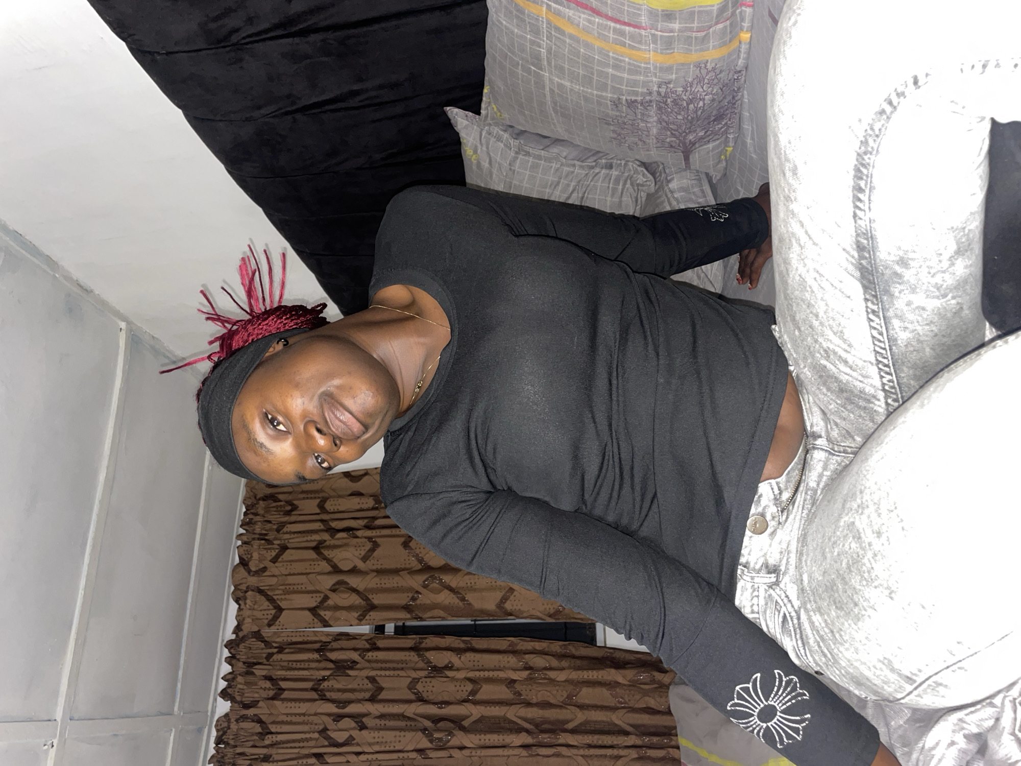 The image depicts an indoor scene in what appears to be a residential setting, located in the city of Jos, Nigeria. A young adult female with dark skin and red braided hair, partially covered by a black headband, is seated on a bed. She wears a black long-sleeved ribbed shirt with a white floral design on the left forearm and light grey acid-wash jeans, the zipper of which is partially unfastened, revealing a silver button with "SHINE" inscribed. A thin gold chain necklace and a small gold hoop earring are visible. She looks directly towards the viewer with a slight smile. The bed is made with light-colored sheets and features two pillows with a grey grid pattern, accented with yellow, pink, and purple lines, and a dark grey tree-like motif. Behind her to the right is a dark, possibly upholstered, headboard. The background on the left shows dark brown curtains with a repeating geometric pattern, partially revealing a window. Above, a white or light grey ceiling with exposed wooden joists is visible. Her right arm is bent, with the hand resting on the bed, while her left arm rests across her lap.
