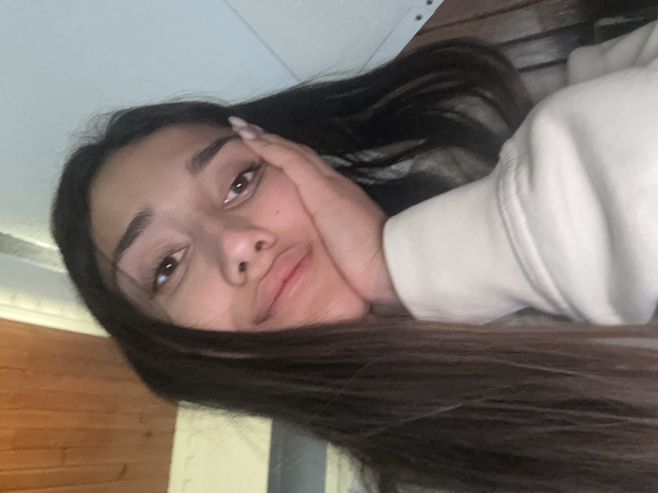 A close-up, slightly blurry image depicts a young female individual indoors in the city of Tutin Municipality, Serbia. She has long, dark brown hair, fair skin, and is looking towards the viewer with a slight smile, her lips partially pursed. Her left hand props her chin and lower jaw, with the fingers resting against her cheek. Visible fingernails are manicured and appear to have a light-colored polish. She is wearing a light-colored, possibly off-white, long-sleeved garment, identifiable by the cuff of the sleeve. The background consists of a light brown, possibly wooden, textured surface in the upper left, transitioning to a light blue-grey painted surface on the upper right and center right. A darker brown or wooden element is visible on the far right. The lighting is soft and even.