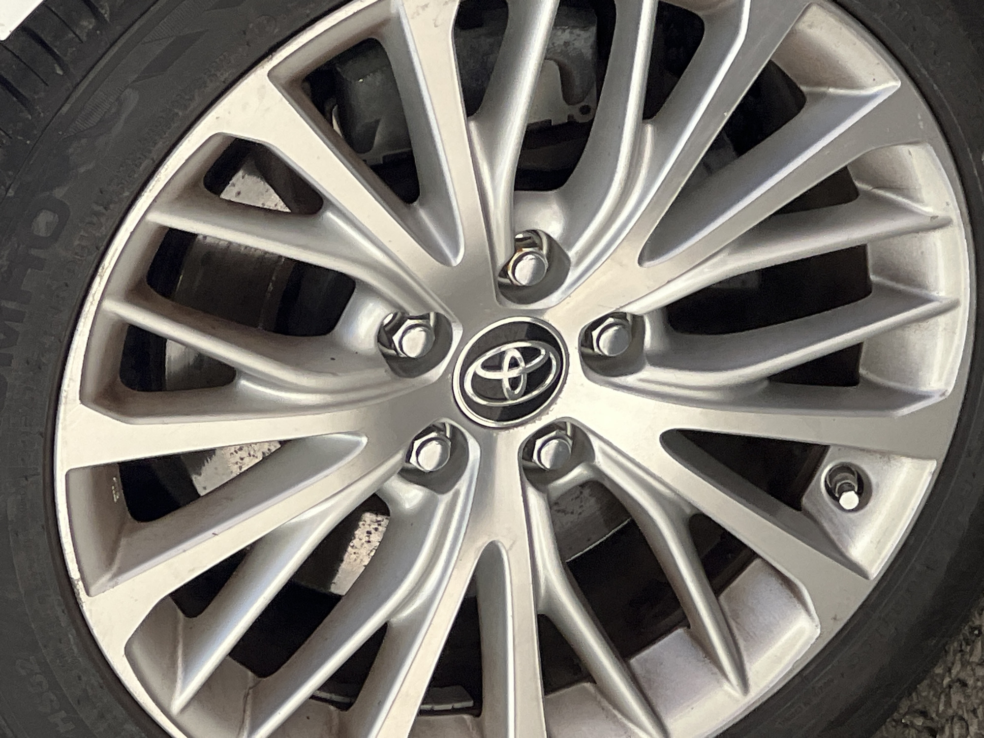 A close-up, eye-level shot showcases the front passenger-side wheel of a silver Toyota car, parked outdoors on an asphalt surface, likely in the city of Donegal, Ireland. The wheel, a 17-inch alloy rim with a multi-spoke design, displays a Toyota emblem at its center. Visible behind the spokes are the car's brake rotor and caliper. The tire is a black Michelin Latitude model, size 225/65R17. No people are present in the image, and no actions are being performed. The environment appears to be a daytime street scene, with no discernible weather conditions.