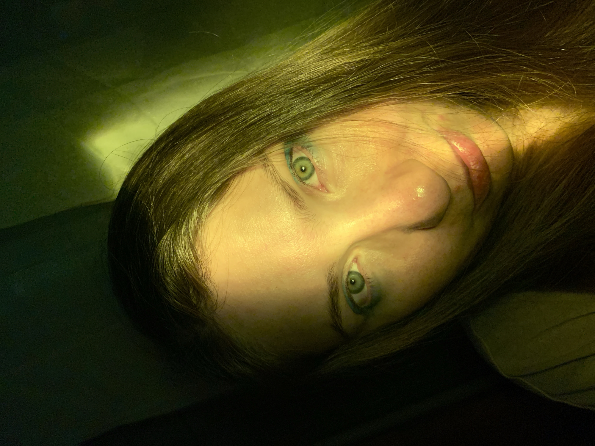 A close-up, low-angle, indoor portrait of a young Caucasian woman is presented. She has long, dark brown hair that frames her face, and her skin appears fair. Her eyes are green and are accentuated with blue eyeshadow. The lighting in the image is dramatic, with a strong yellow-green cast, and a bright light source is visible in the upper right quadrant, casting a harsh shadow on the left side of her face. The background is dark and indistinct, suggesting an indoor setting with controlled lighting. The woman is looking directly at the camera with a neutral expression, her lips slightly parted. There is no indication of the specific location within Chaikovsky, Russia, in this image; it depicts a personal portrait rather than a landmark or a scene identifiable with a particular place.