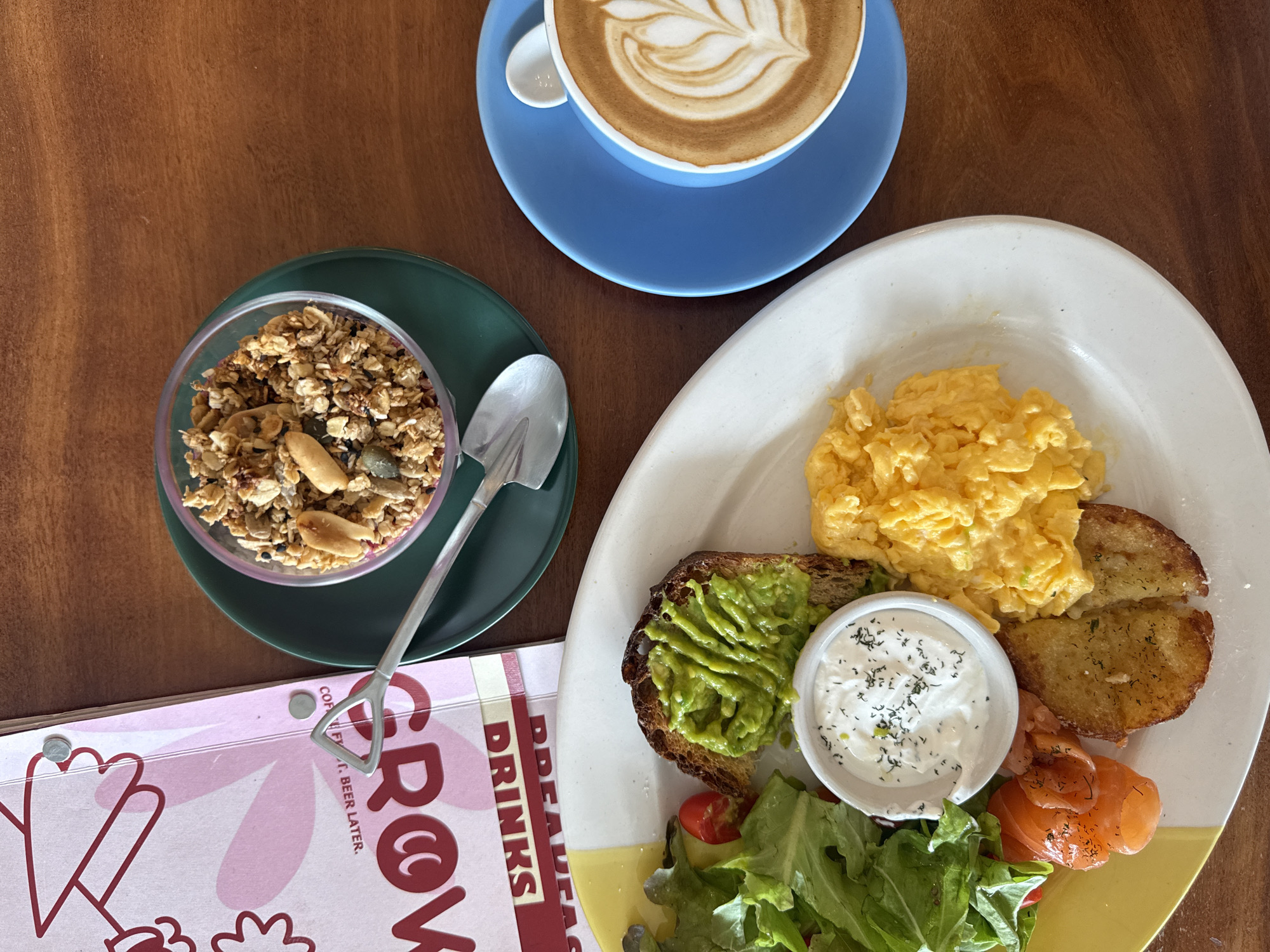 An overhead shot shows a breakfast spread on a wooden table in Ko Samui District, Thailand. A white plate holds scrambled eggs, a piece of toast with mashed avocado, smoked salmon, a small bowl of dip with dill, a small salad, and two pieces of fried potato. To the right, a blue saucer holds a cup of latte art coffee. Above the plate, a glass bowl filled with granola and a small spoon are on a green saucer. A pink menu with the word "GROW" in large red letters is to the left.