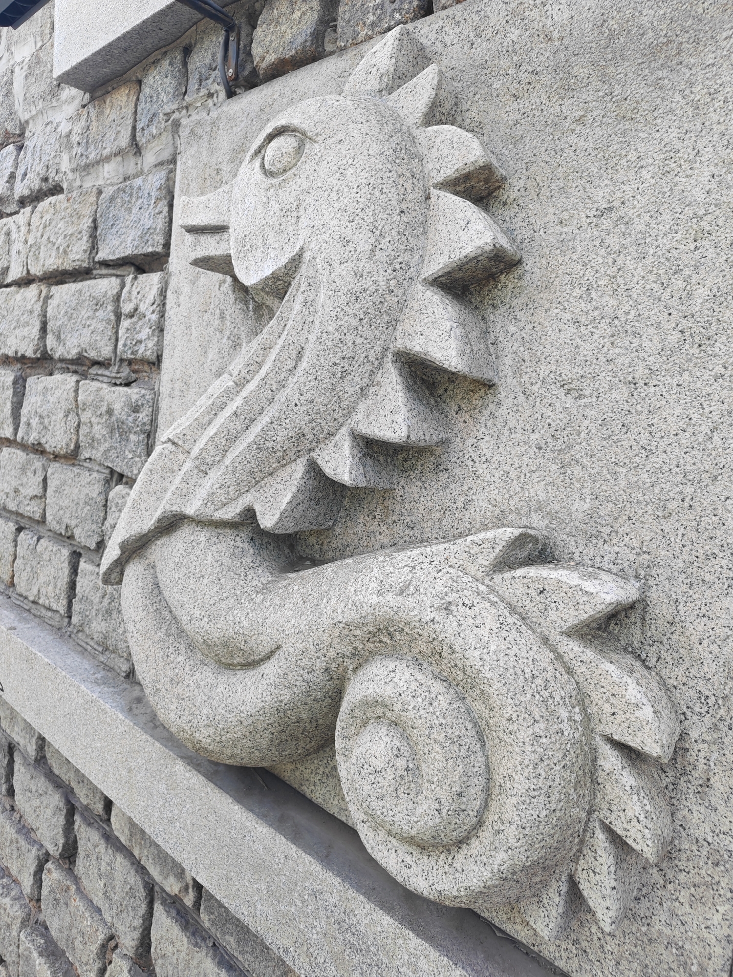 This image showcases a detailed stone carving of a stylized creature, possibly a dragon or a griffin, mounted on a granite wall. The creature is depicted in profile, with a prominent eye, a beak-like snout, and layered, fin-like protrusions along its back and tail. Its body is coiled, suggesting movement or repose. The carving is textured, with the rough, speckled surface of the granite adding to its tactile quality. The background consists of a rough-hewn stone wall, composed of rectangular blocks, with a darker, possibly metal, fixture attached to the upper portion of the wall, hinting at the surrounding architecture. A smooth, projecting stone ledge runs horizontally in front of the carving. The overall impression is one of permanence and artistic craftsmanship. The lighting suggests it is daytime, with the sunlight creating subtle shadows that highlight the three-dimensional relief of the sculpture. The location is identified as Vladivostok, Russia. There is no visible text in the image.