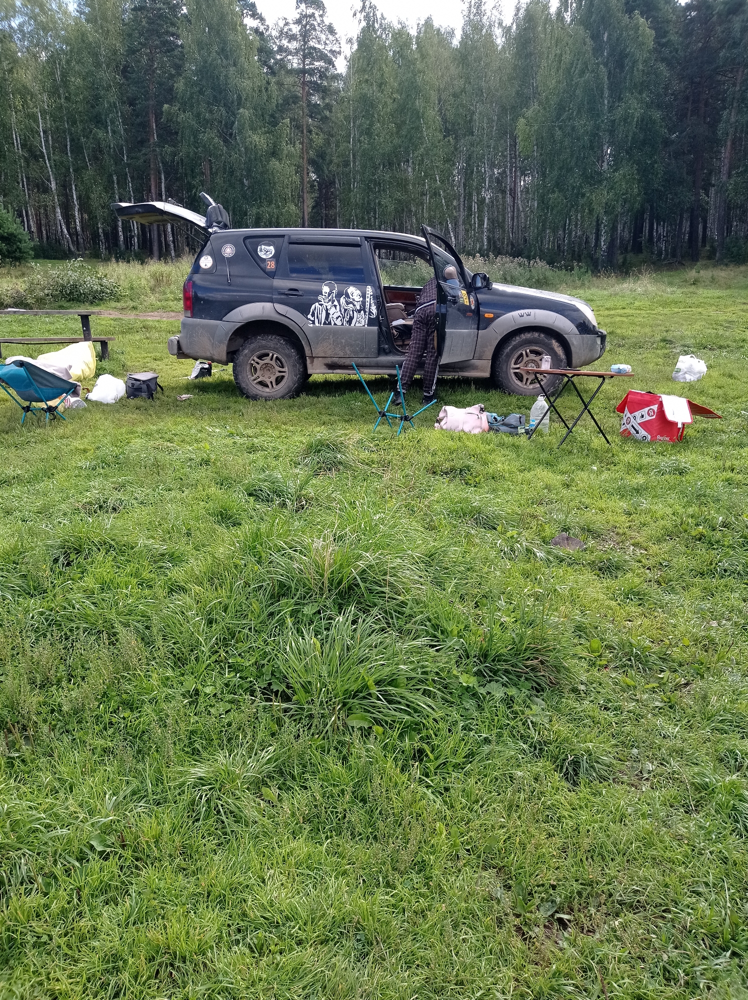 A dark-colored SUV with its rear hatch and driver's door open is parked in a grassy field near a forest in Degtyarsk, Russia. The SUV has several decals on its side, including a number "28" and what appear to be graphic stickers of people. A person in checkered pants is standing in the open driver's door, partially inside the vehicle. Various items are scattered around the vehicle on the grass, including a red cooler, a water bottle, a pink bag, a black bag, and two camping chairs. The environment is a natural setting with tall grass and a dense forest in the background. The sky is overcast.