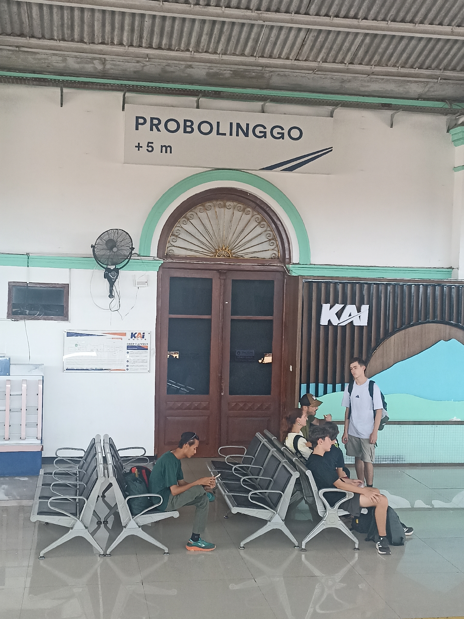The image depicts the interior of a train station waiting area in Probolinggo, Indonesia. A sign overhead reads "PROBOLINGGO +5 m". Several people are seated on metal benches. On the left, a man in a green shirt is looking at his phone. To his right, a group of four people, two women and two men, are conversing. Further to the right, a young man with a backpack is standing. The environment includes a large wooden door, a wall-mounted fan, and a KAI (Indonesian Railway Company) logo. The floor is tiled and reflects the lighting. The overall atmosphere is that of a typical public waiting area.