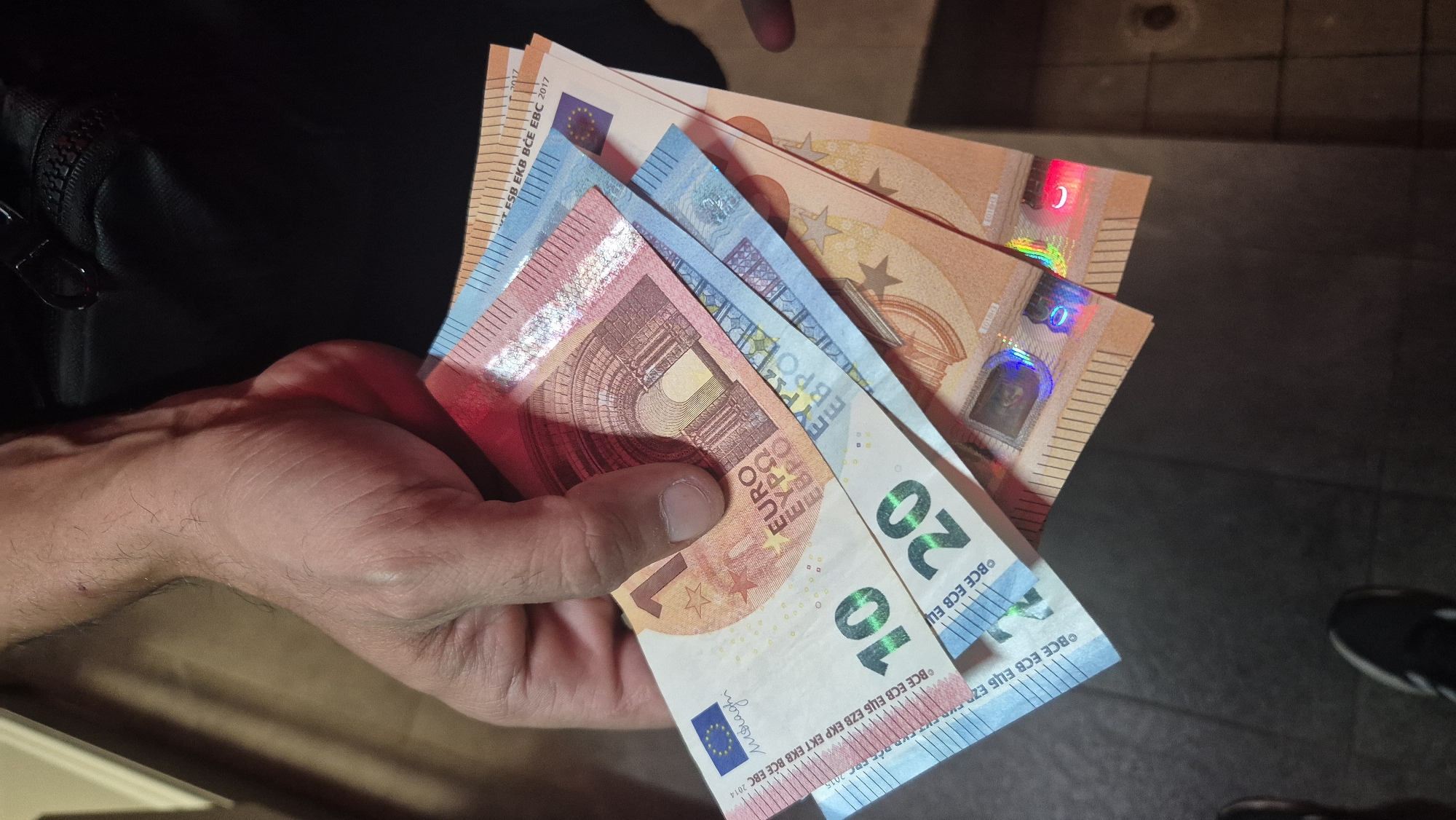 A hand holds several Euro banknotes, fanned out to reveal their denominations. The visible denominations are a 10 Euro note, a 20 Euro note, and parts of what appear to be a 50 Euro note and possibly another note further back. The banknotes are of different colors and designs, consistent with European currency. The background is dark and appears to be indoors, with some hints of light reflections on the banknotes suggesting artificial lighting. The image does not contain any specific visual cues that would indicate its location in Lovech, Bulgaria, or any other city.