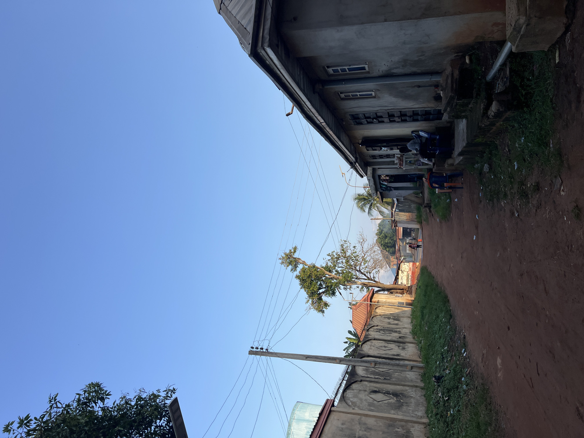 The image depicts a street scene in Nkpor, Nigeria. The foreground is dominated by a red dirt road. To the left of the road is a high, textured concrete wall with a barbed wire topping, separating it from adjacent properties. To the right, a long, one-story building with a gray, stucco-like exterior lines the road. This building has several windows with bars, and a sloped, dark metal roof. In front of this building, there is a raised concrete walkway or foundation.
Further down the road, several individuals are visible. Two people are sitting on chairs outside the building on the right, and another person is walking away from the camera further down the road, carrying an orange bag. A few more figures and structures are faintly visible in the distance, suggesting a populated area. A large tree with sparse foliage stands in the middle ground, near the wall on the left. Electrical poles and wires are prominent throughout the scene, indicating infrastructure. The sky is clear and bright blue, suggesting it is daytime. The overall impression is that of a typical, modest street in a Nigerian town.
