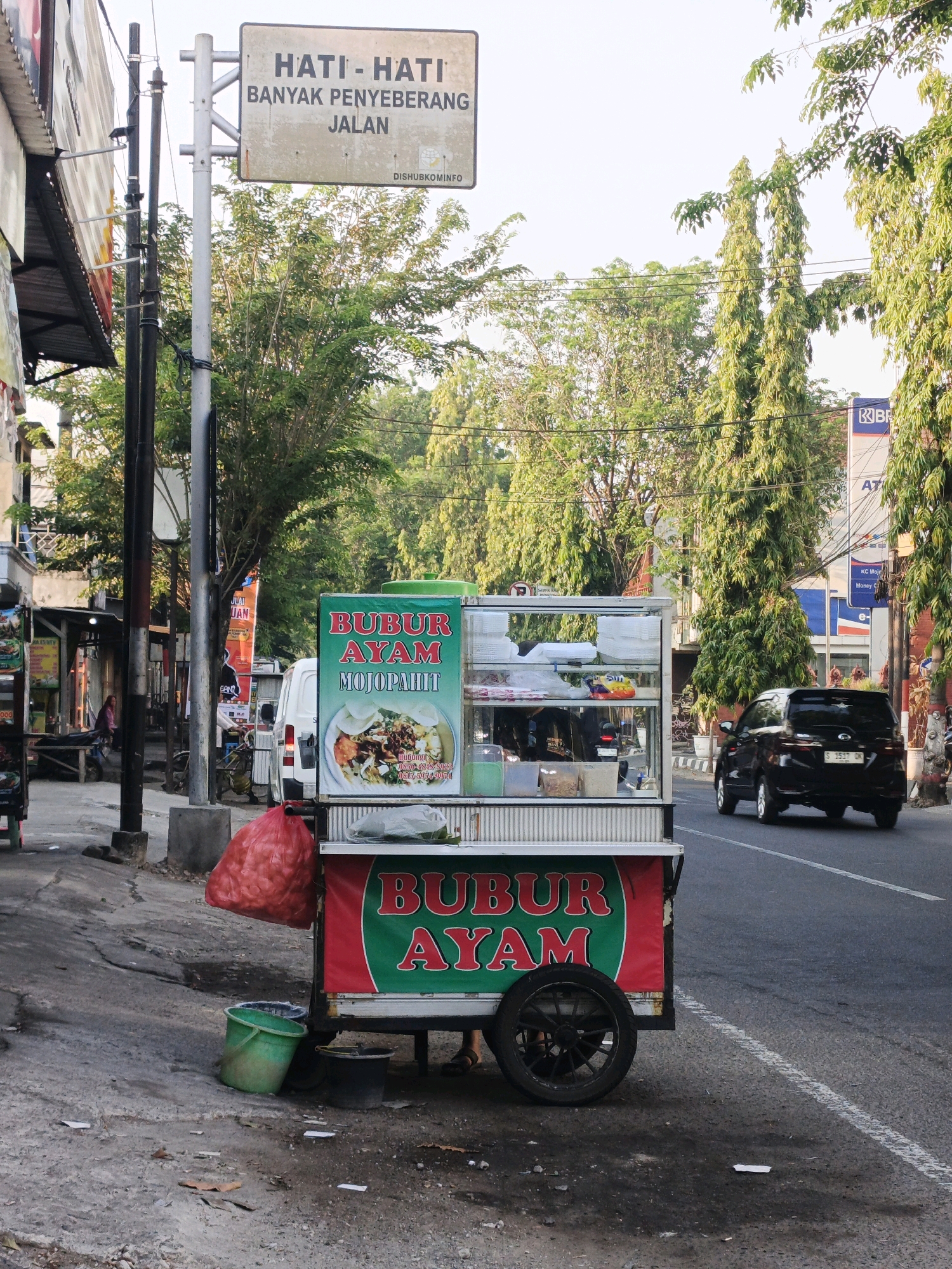 The image depicts a street scene in Miji, Indonesia, likely during daylight. A food cart is prominently positioned on the side of a road, displaying signage that reads "BUBUR AYAM MOJOPAHIT". The cart is laden with food items and is equipped with a glass display case. A large red plastic bag, filled with what appears to be onions, is tied to the side of the cart. Two green and black buckets are on the ground next to the cart.
In the background, a black SUV is driving on the road. A white van is parked behind the food cart. Several trees line the street, with their branches extending over the road. A traffic sign is visible on a pole, with text in Indonesian that translates to "BE CAREFUL - MANY PEDESTRIANS". Shops and other structures are visible along the left side of the street. The overall environment suggests a busy urban or semi-urban setting with commercial activity.