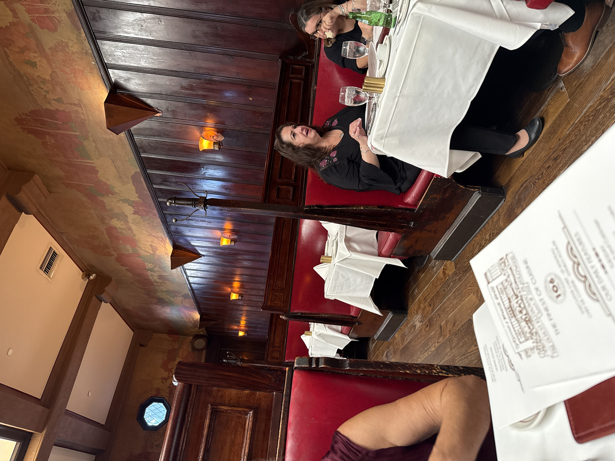 This is a medium shot photo of two women seated at a table in a restaurant. The woman on the left is wearing a black top with floral embroidery and is speaking with her hands gesturing. The woman on the right is wearing glasses and a black top, and is eating. Both women are seated in a booth with red leather upholstery. The table is covered with a white tablecloth and set with water glasses, a bottle of water, and what appears to be a menu. The restaurant's interior features dark wood paneling, a mural on the upper wall, and ornate light fixtures. The floor is made of wood planks. The scene suggests a dining experience in a well-established or classic restaurant, possibly in Los Angeles, United States of America, known for its traditional ambiance. The photo captures a moment of conversation and dining.