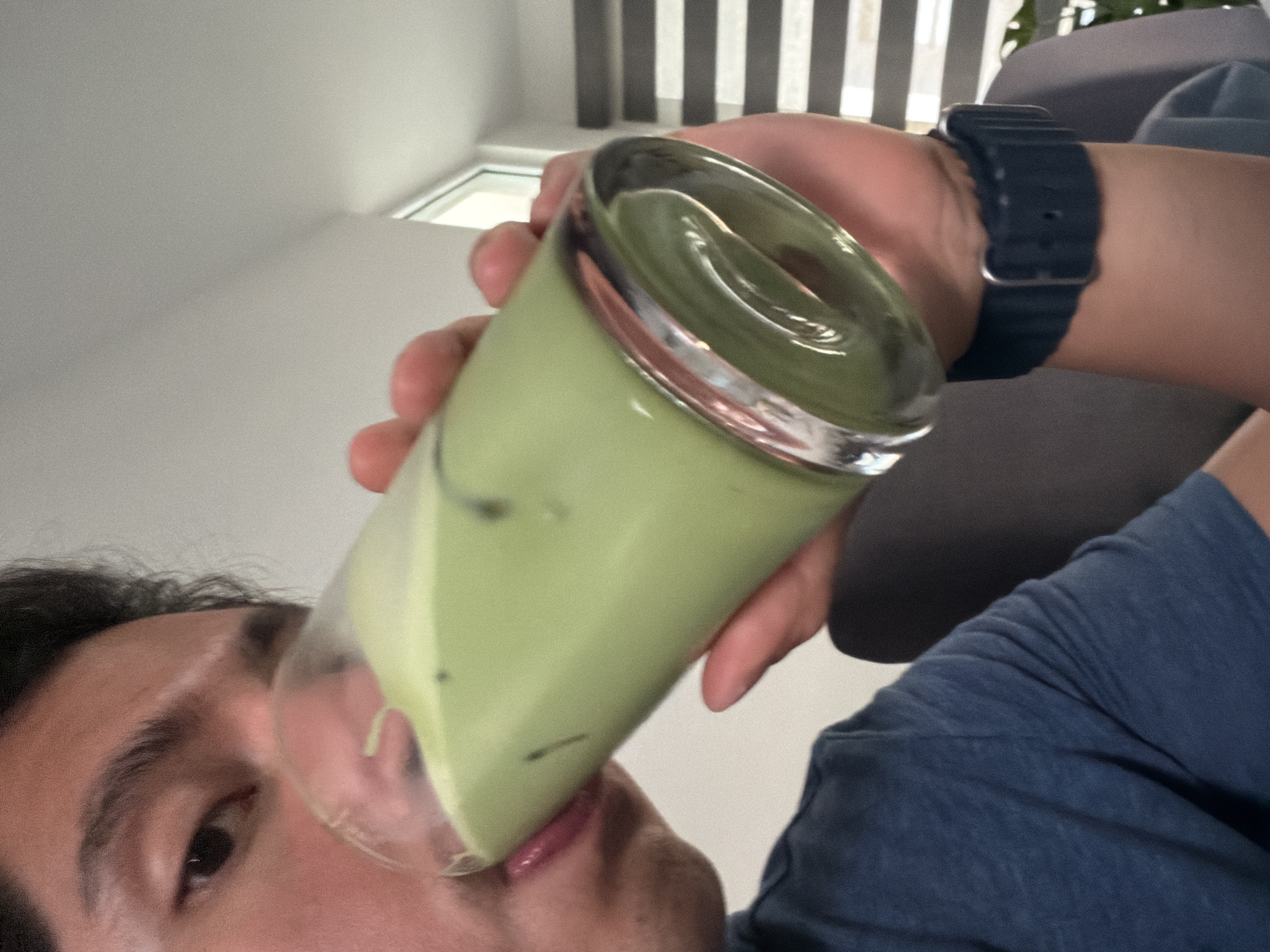 A person is drinking from a glass filled with a light green beverage. The person is wearing a dark blue t-shirt and a dark blue smartwatch. The background features white blinds and a grey couch. There is no indication that this image was taken in Santa Clara Ocoyucan, Mexico.