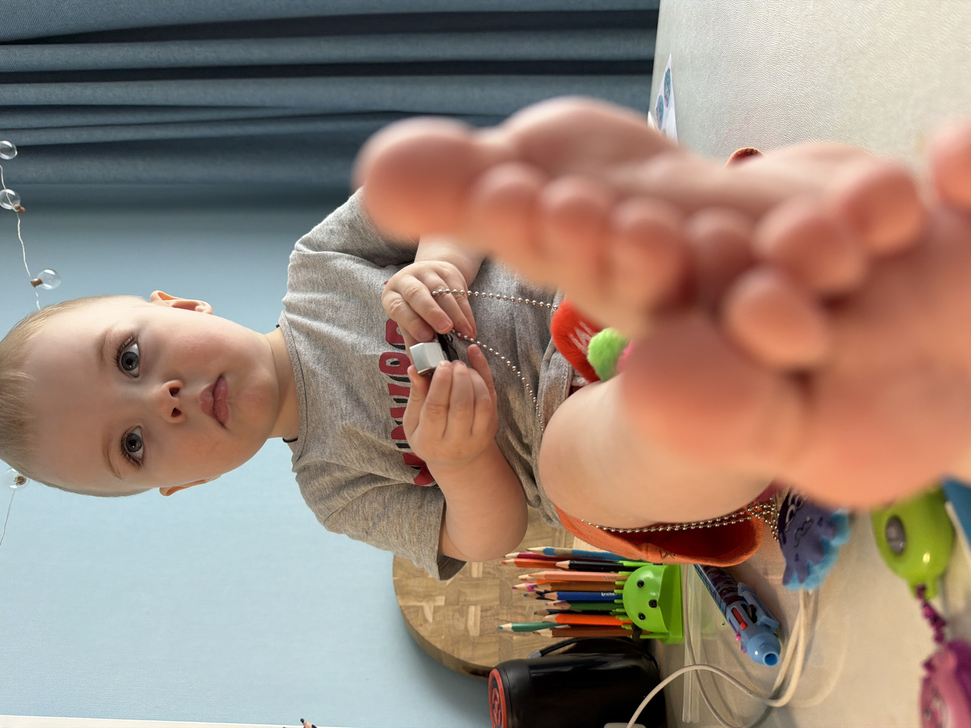 A toddler is holding a small silver rectangular object with a beaded chain in his hands. A group of colored pencils are held in a green pencil holder on a white surface with a wooden cutting board and black and red pencil sharpener. The toddler is wearing a gray t-shirt. A beaded string of lights hangs in the background against a blue wall. One of the toddler's feet is in the foreground. It is not possible to determine the location in Votkinsk, Russia from the image.