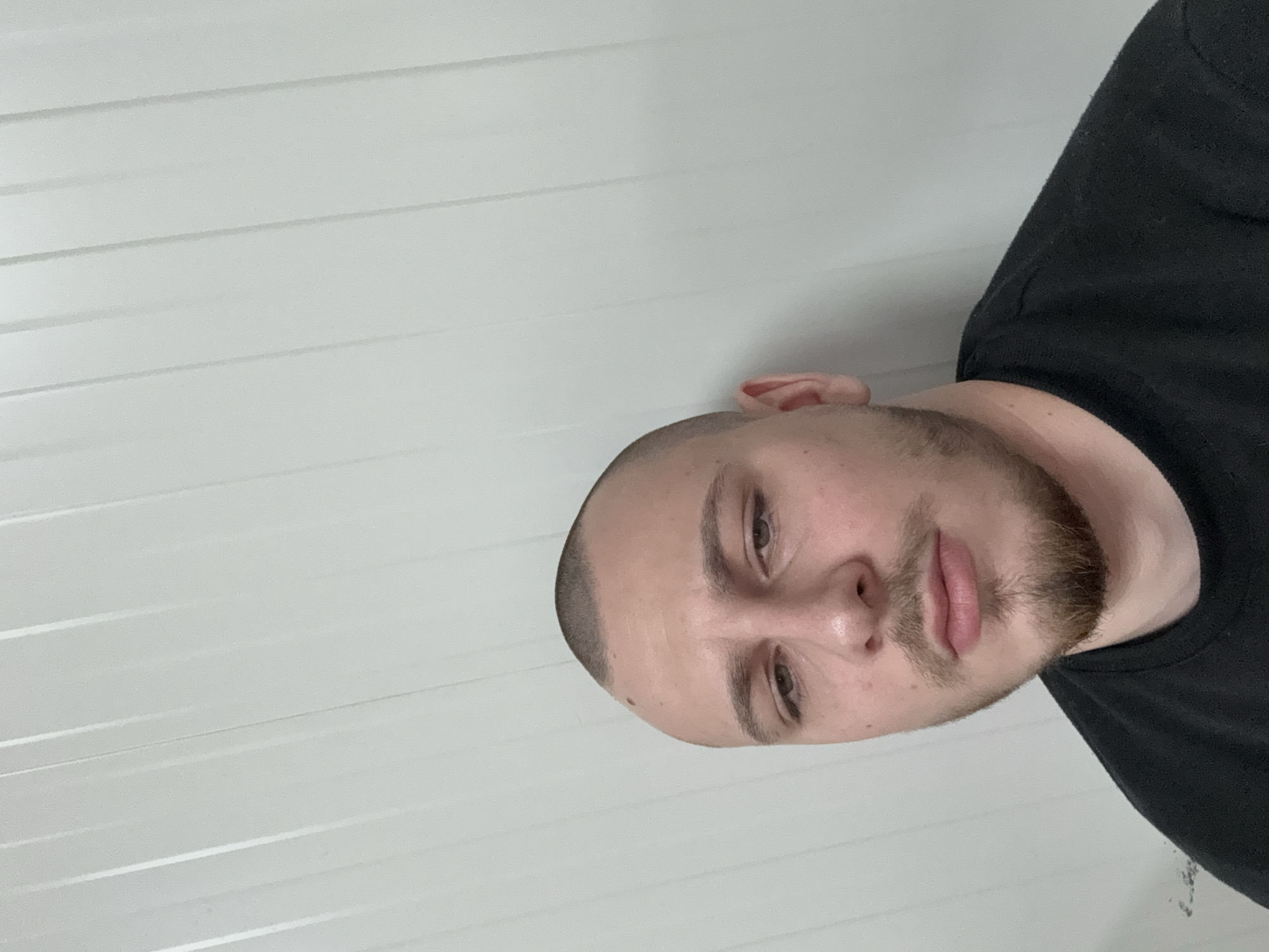 A close-up shot depicts a man with a close-cropped haircut and a short, well-trimmed beard. He wears a black shirt. The background consists of a white, horizontally-paneled wall. There is no evidence to suggest the man or the wall is located in Stara Zagora, Bulgaria. The image provides no additional contextual information.