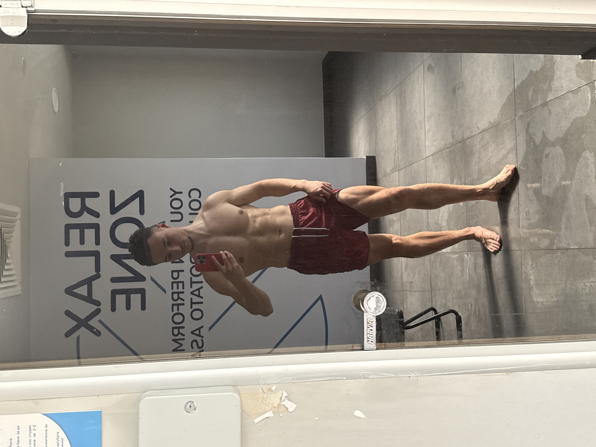 A bare-chested man in maroon shorts is taking a picture of himself in a mirror. The mirror is affixed to a wall with writing on it, reading "RELAX ZONE," and in reverse "JOY" and "PERFORM". Below the writing is "АГА OTATOS". The man holds a red phone in his left hand. His right arm is held at his side. The background is a gray tiled wall and floor. There is a black metal object, possibly a towel rack, behind the man. Lower left, there is a white plate covering something, along with peeling paint or stickers. In front of it is a blue sign in Cyrillic. There is a metal Harvia sticker on the glass. Based on the Cyrillic text, the image may be taken in Bulgaria, specifically Sofia.