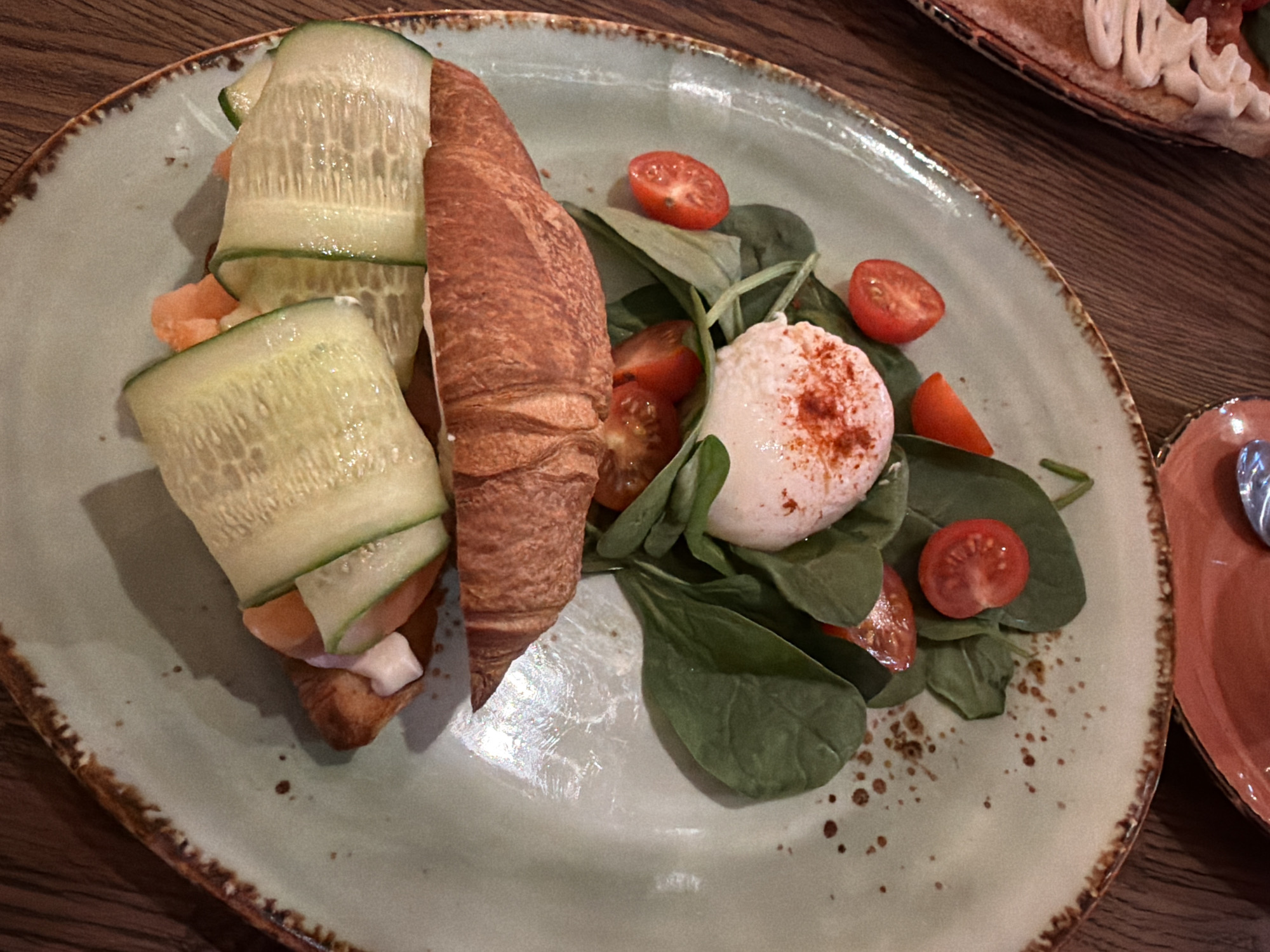 The image depicts a plate of food. The plate is light green with a brown rim and contains a croissant cut lengthwise, filled with salmon and topped with cucumber slices. Next to the croissant is a salad of spinach leaves, cherry tomatoes, and a poached egg dusted with red pepper flakes. The plate sits on a wooden table. In the background, there is another plate with a pastry topped with white frosting. A small orange plate with a spoon is partially visible on the right side of the image. There is no information regarding the location in the city of Chaikovsky, Russia.