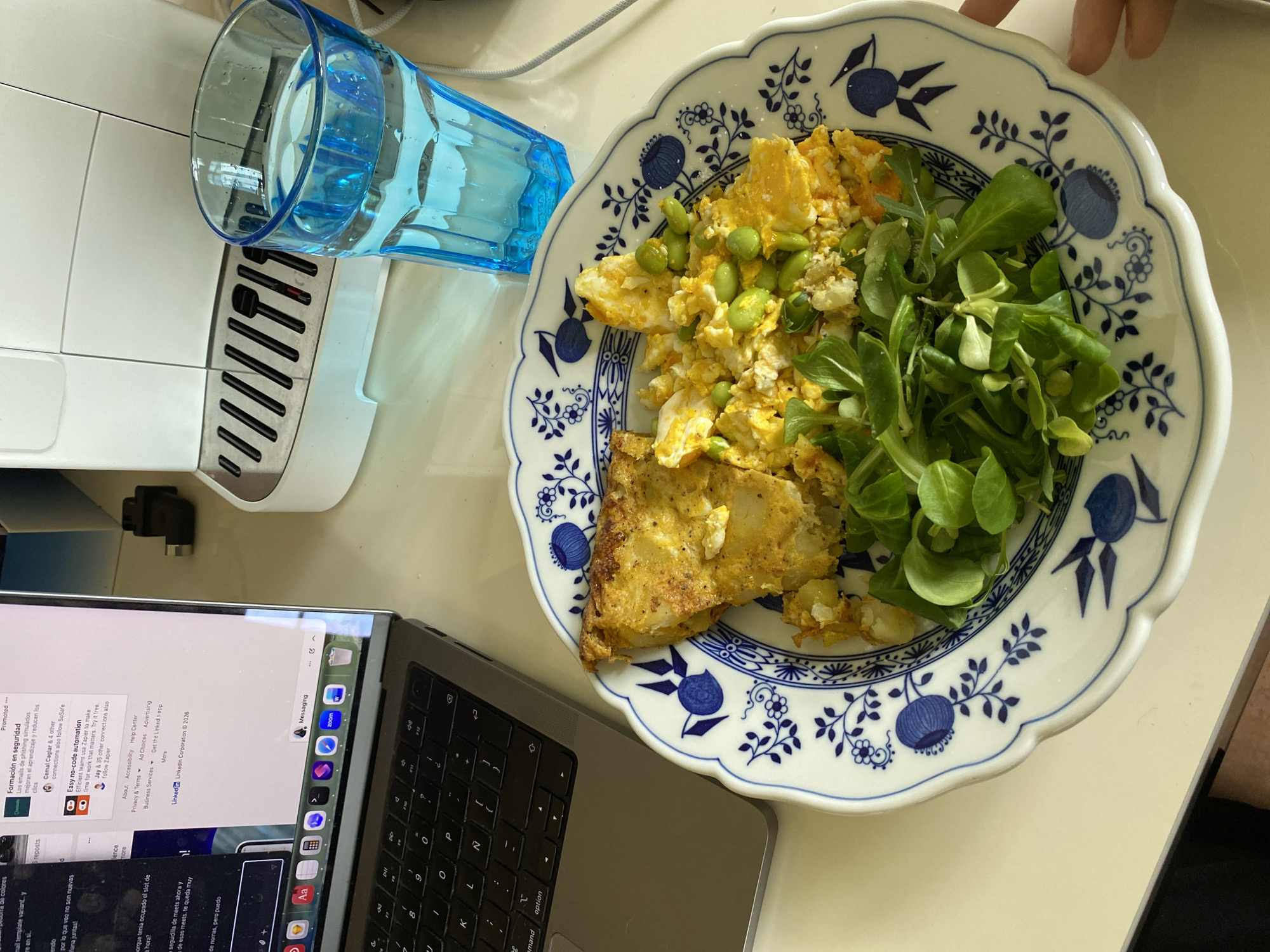 The image shows a close-up, elevated view of a cluttered workspace. A laptop is on the left side, displaying a web browser with various windows and tabs open, including LinkedIn and messaging applications. To the right of the laptop, a plate with a blue and white floral design holds a meal consisting of what appears to be a slice of quiche, scrambled eggs with edamame, and a side of greens. A blue glass filled with water is next to the plate. A white appliance, potentially a coffee maker, is visible in the upper left corner. The scene suggests someone is eating a meal while working on their laptop. There is no information provided that indicates that this location is in Villaviciosa, Spain.