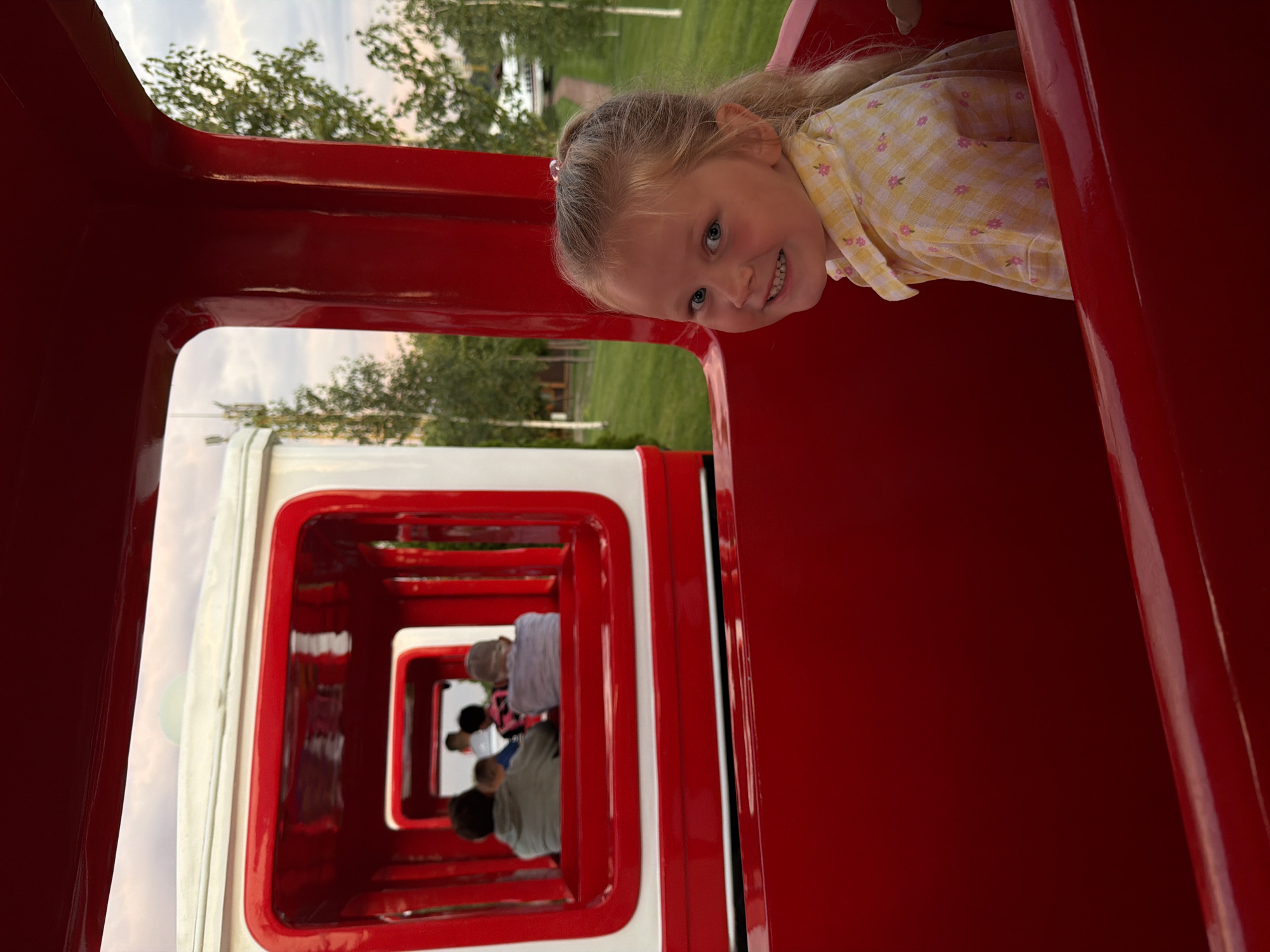 In the city of Kamyshin, Russia, a young girl with blonde hair pulled into a ponytail and blue eyes smiles at the camera while sitting in a red train car. She is wearing a yellow checkered shirt with pink floral accents. Behind her, other people are visible in the train, seated in a row. The train appears to be on a track outdoors, with a grassy area and trees in the background, suggesting it might be a park or amusement area. The sky above is overcast. The perspective is from inside the train, looking towards the back and then to the side.
