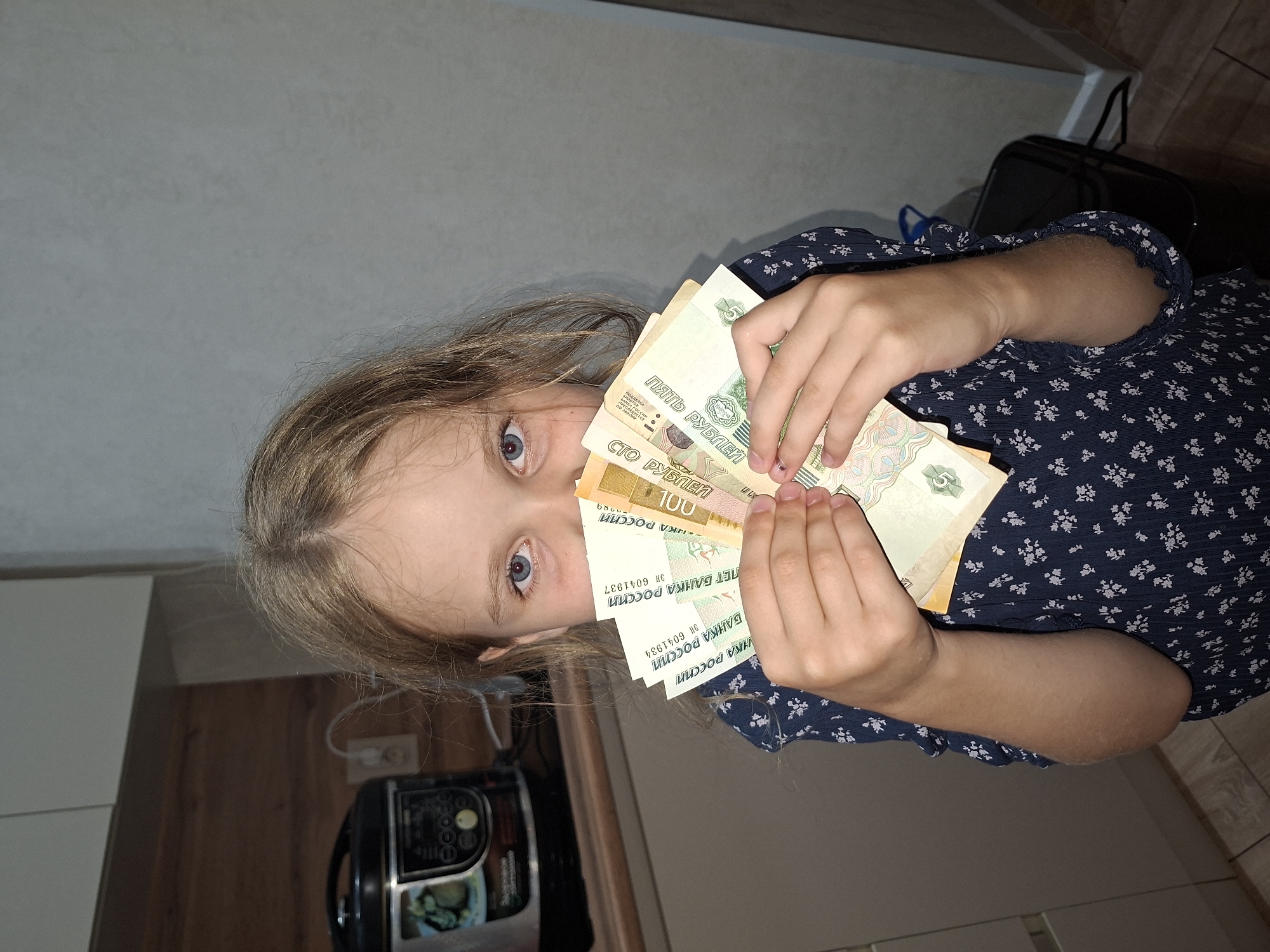 A young girl with light brown hair and blue eyes holds several Russian rubles in front of her face, obscuring her mouth. She is wearing a dark blue dress with a white floral pattern. The background appears to be a kitchen, with kitchen cabinets and a black appliance, possibly a multicooker, visible on a countertop. The lighting suggests an indoor setting, likely during the daytime. The scene does not provide any specific details that would allow for identification of a location within the city of Izhevsk, Russia.