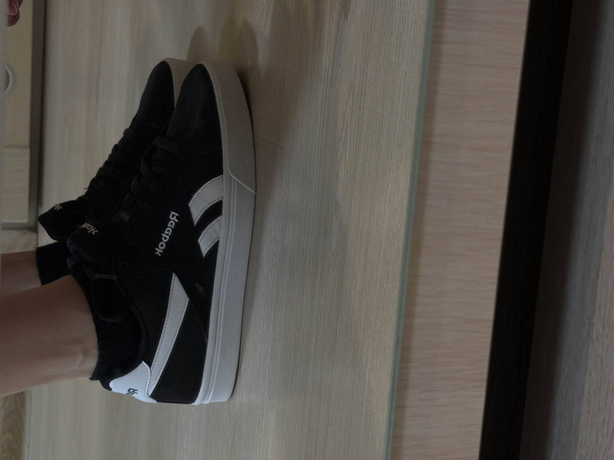 A pair of black Reebok sneakers with white stripes and soles rest on a light wood-grained surface. A person's arm and a portion of their leg are visible, suggesting they are wearing the sneakers. The scene is likely indoors, possibly a room with wooden furniture or paneling. The text "Reebok" is prominently displayed on the side of one shoe, mirrored. The background includes a light wooden surface and a darker wooden panel. The image does not provide information to identify its location as Izhevsk, Russia.
