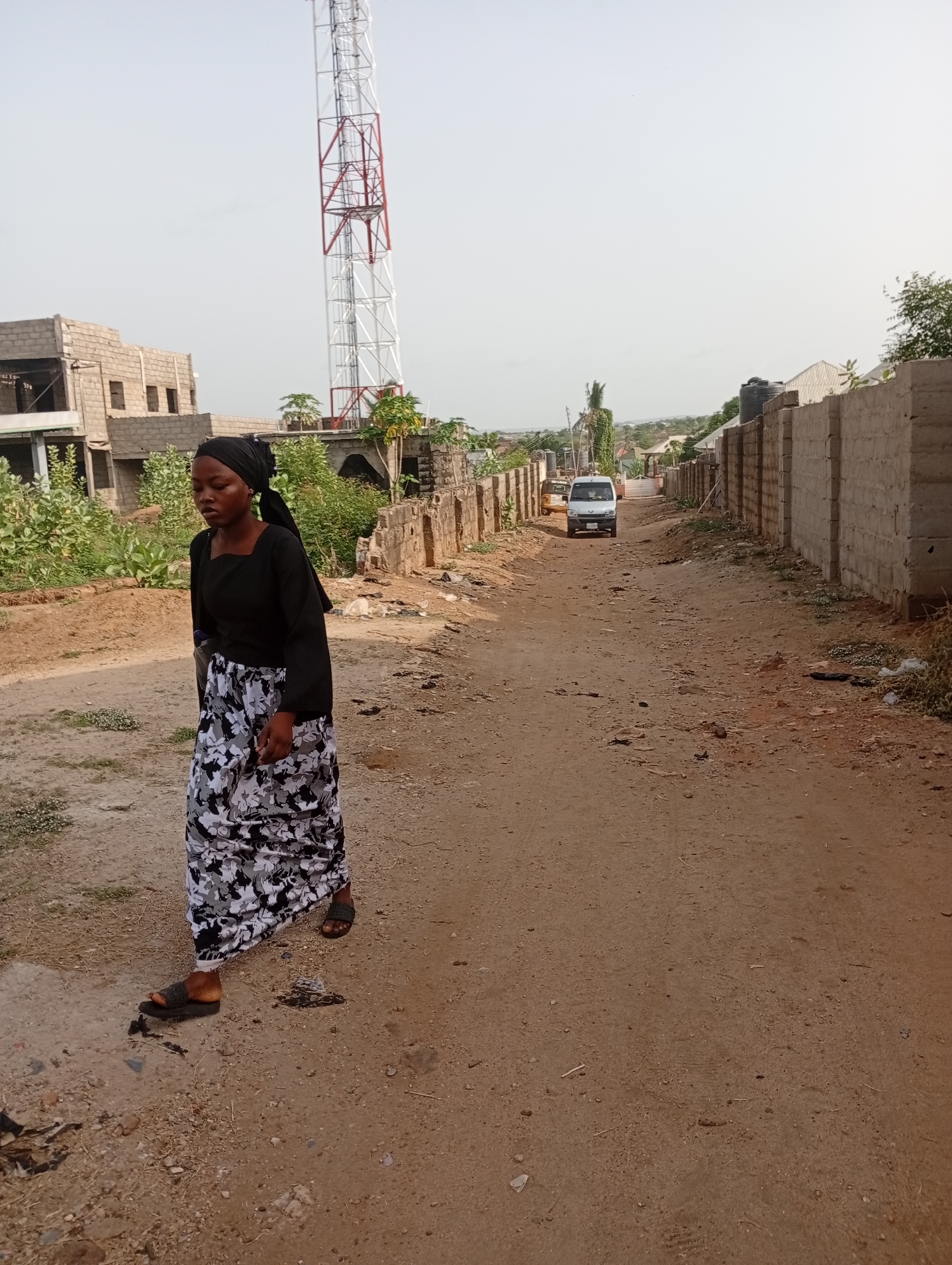 A woman walks on a dirt road in Adankolo, Nigeria. She wears a black top, a long black and white floral skirt, and sandals. She has a black headscarf. To the right, a concrete block wall runs along the road, and to the left, there is a partially constructed building and overgrown vegetation. A white van and a yellow tricycle are visible in the distance. A tall, red and white communication tower rises in the background. The road is dusty and strewn with debris.